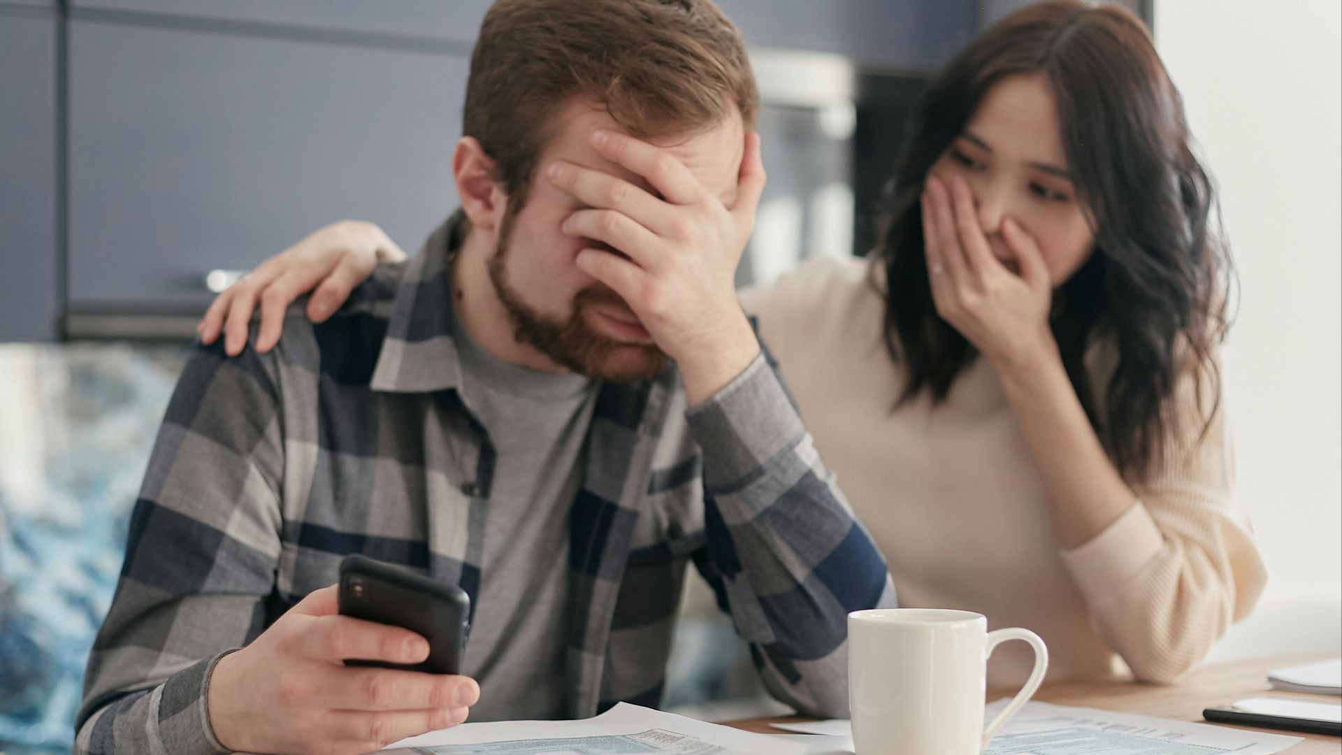 A couple looking stressed and holding a cellphone while reviewing financial documents indoors. Hands on faces in concern.