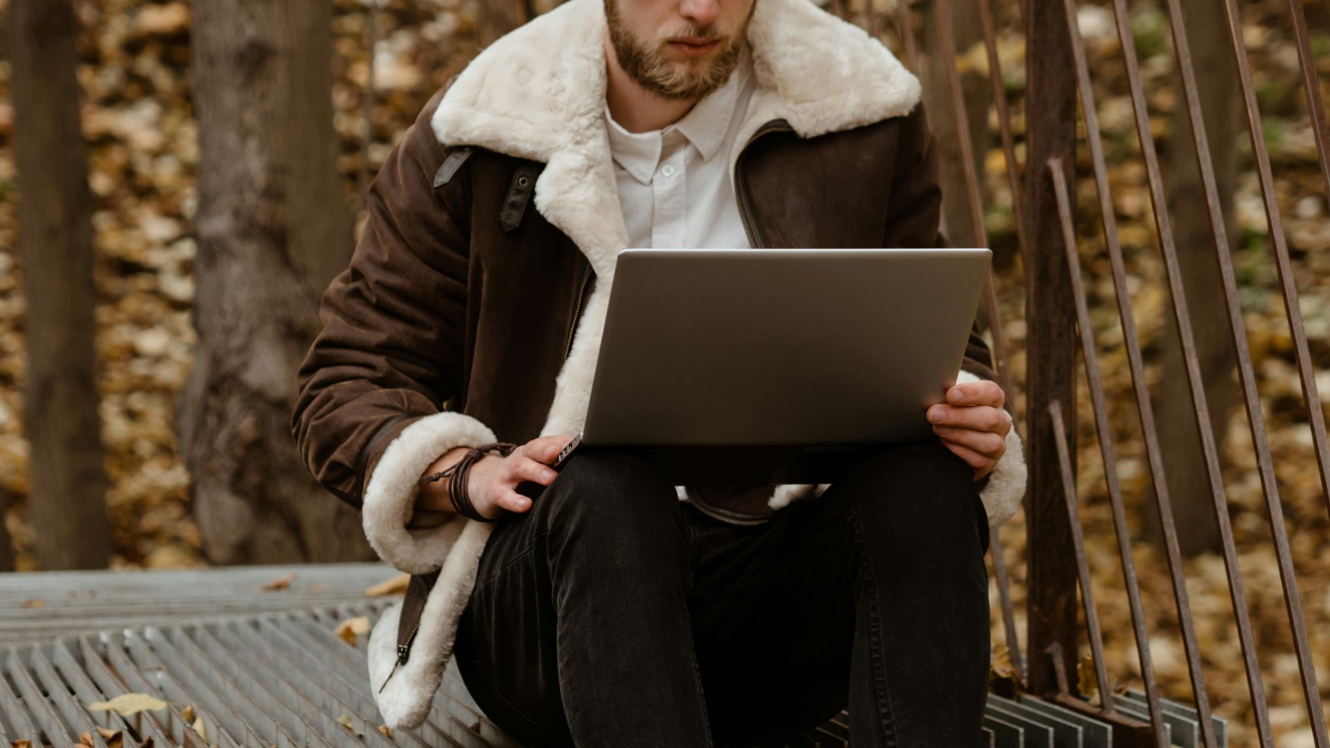 Stylish man working on laptop in an autumn park, embracing freelance lifestyle amidst natural scenery.