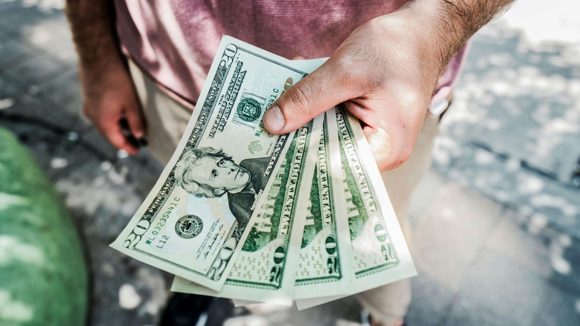 A person holding several twenty-dollar bills outdoors, depicting payment or financial transaction.