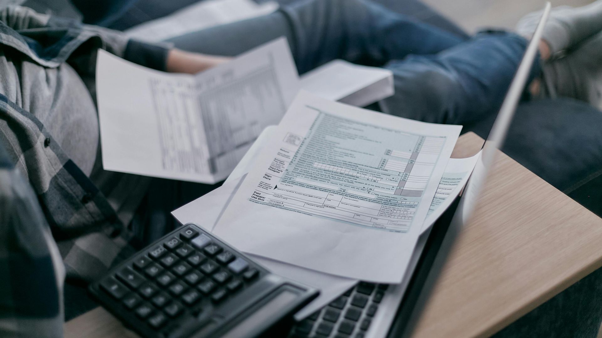A young adult using a calculator and laptop to manage finances at home, surrounded by papers.