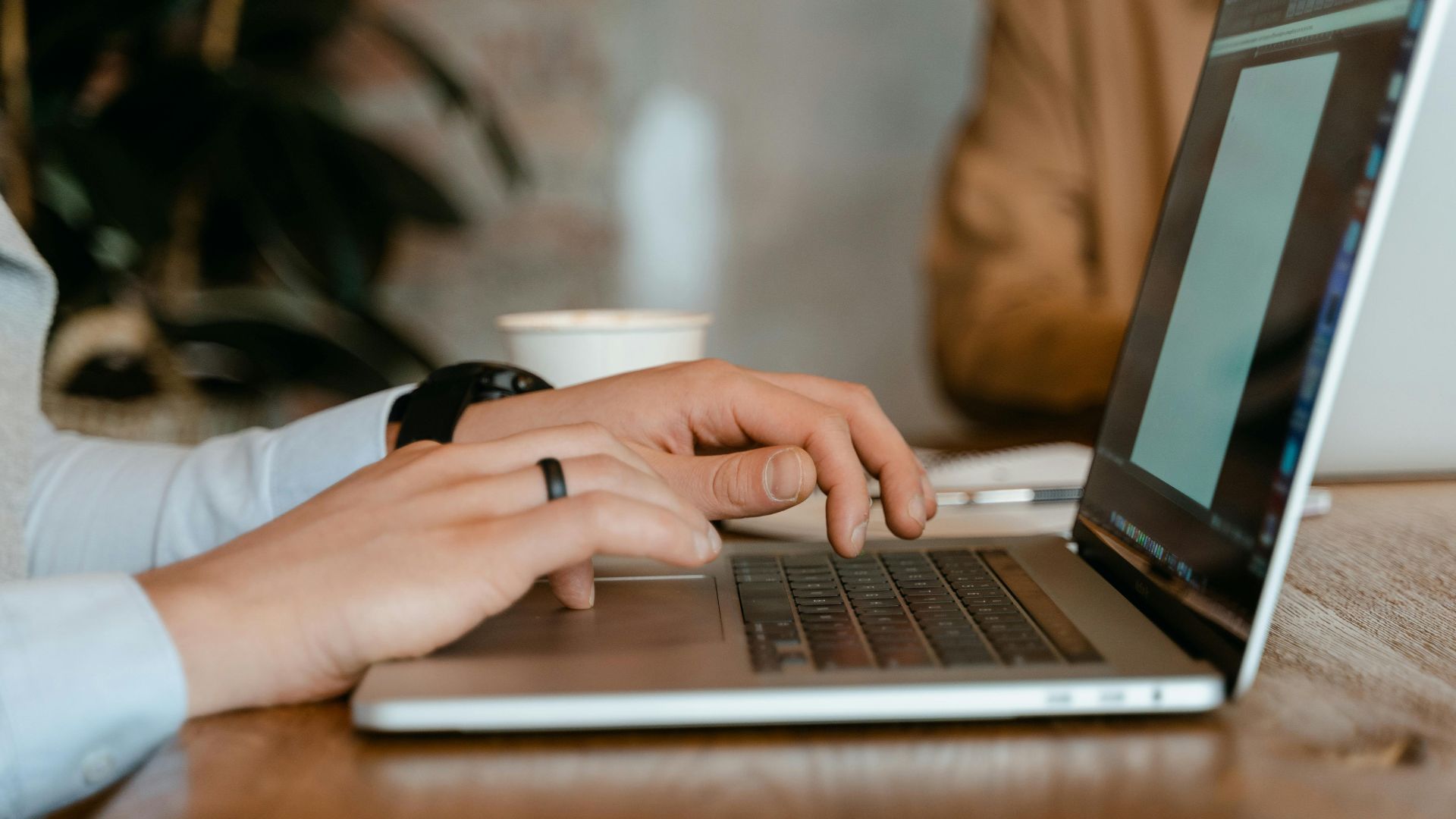 A person typing on a laptop at a wooden table in a modern office setting.