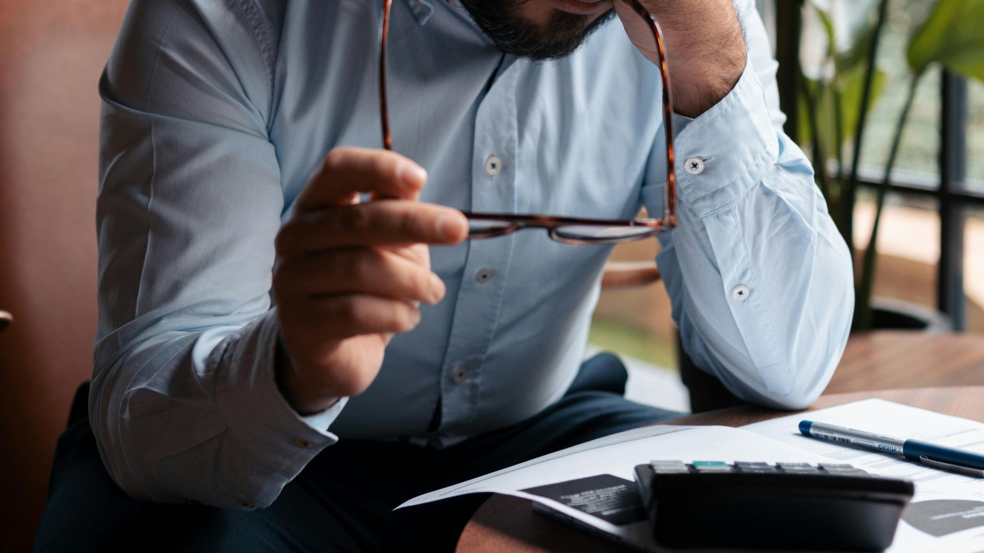 A man sitting at a table, holding his head in stress, facing tax papers and a calculator.