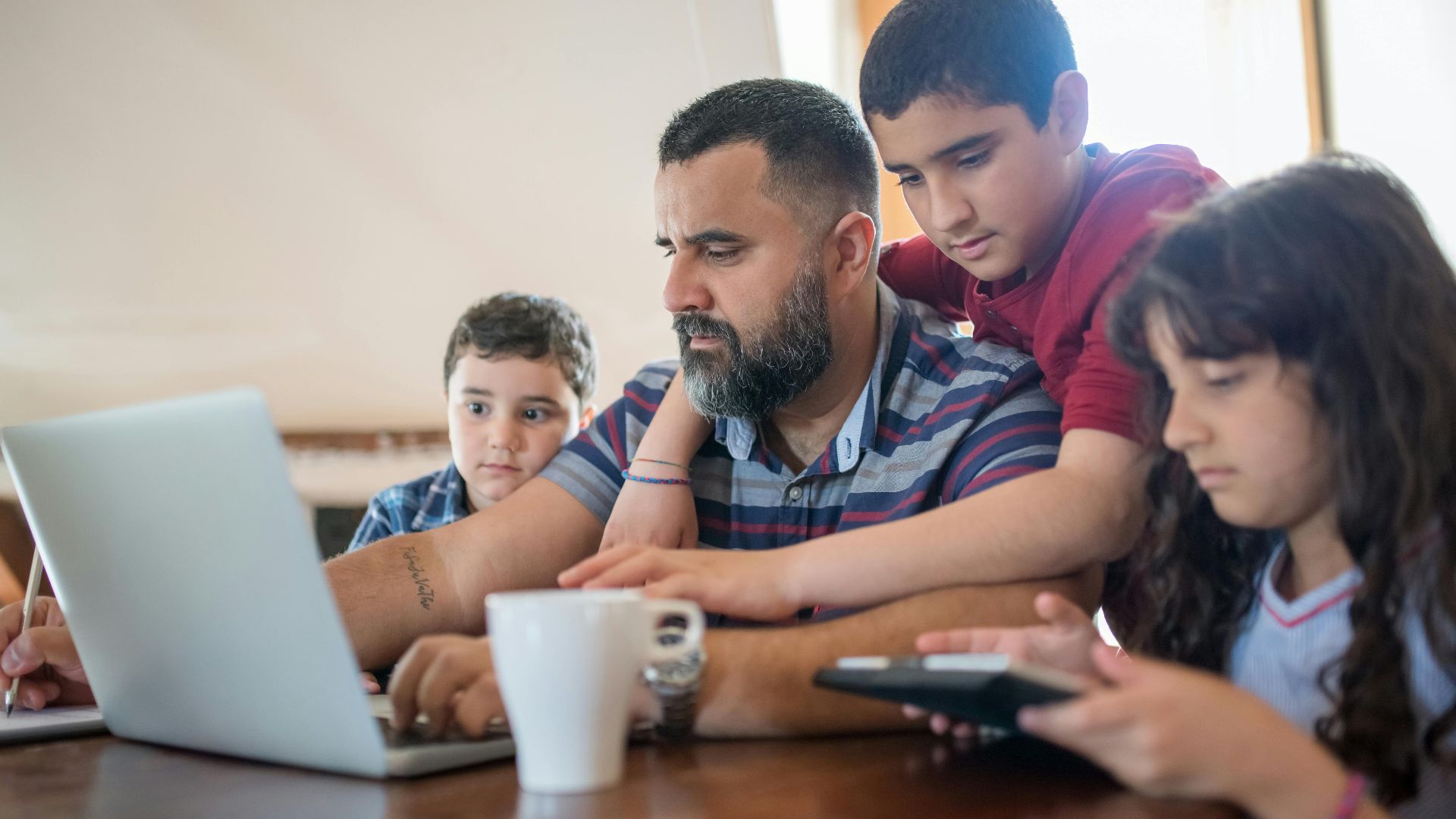 A father manages work tasks at home while bonding with his children around a table.