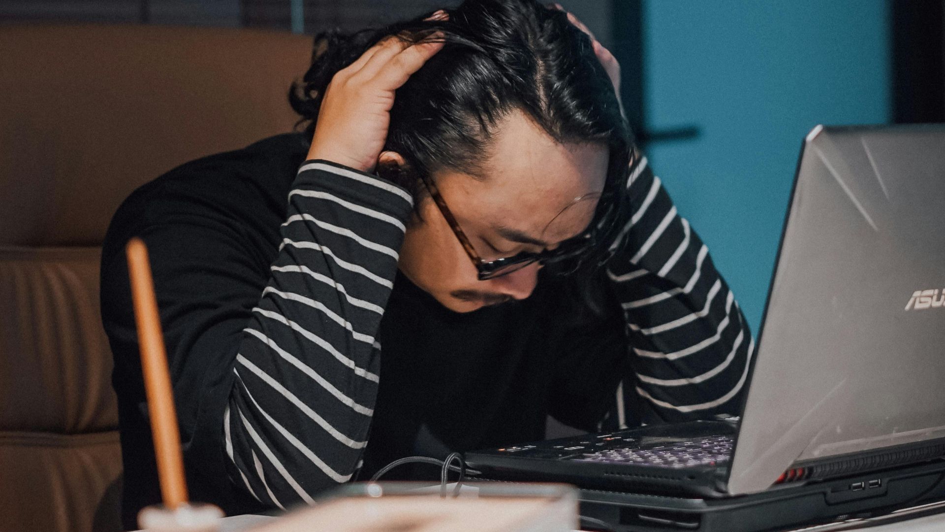 Overwhelmed man working late on laptop in home office, looking stressed and exhausted.
