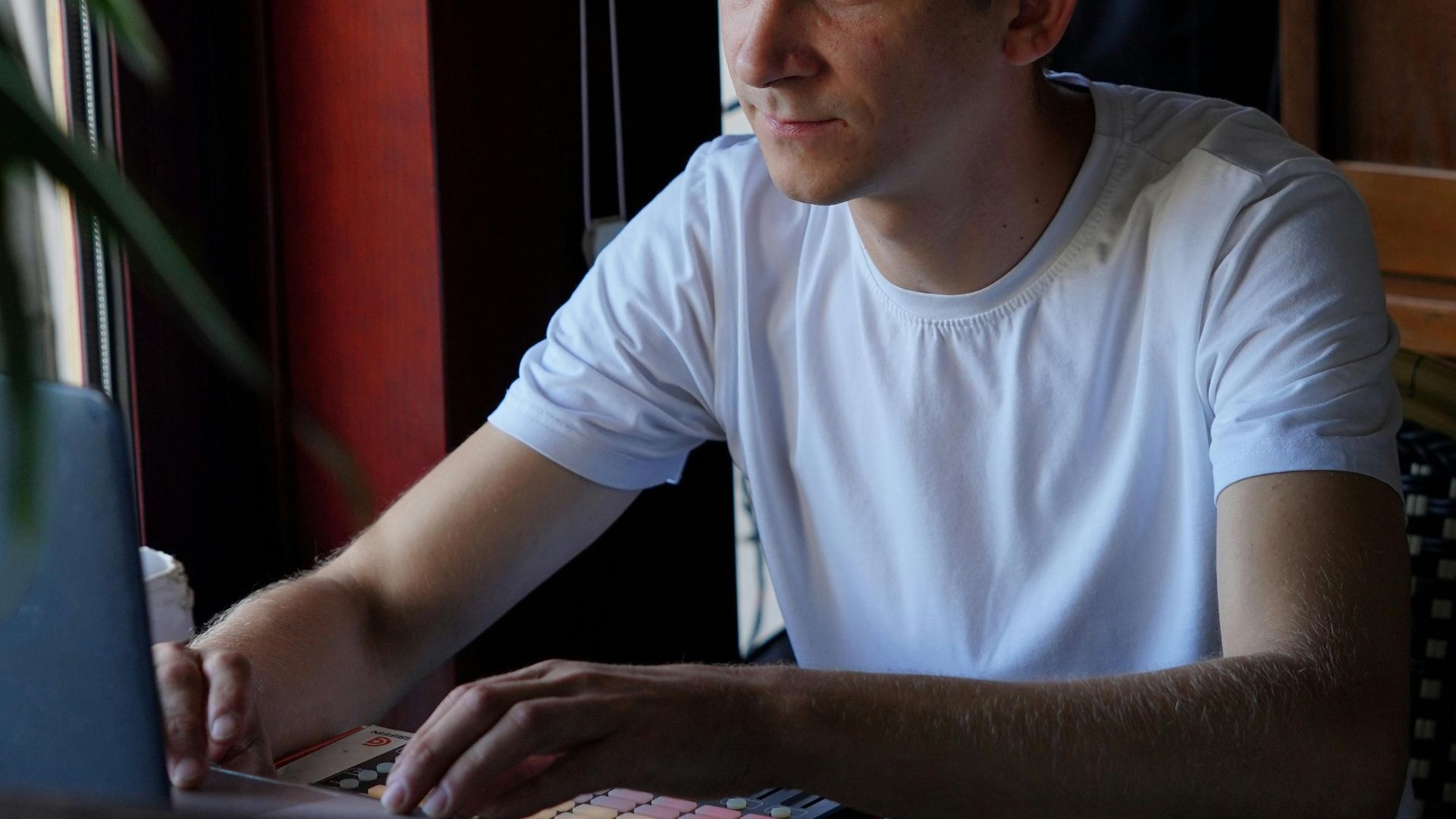 A young man in a café working on his laptop by the window, focused and casual.