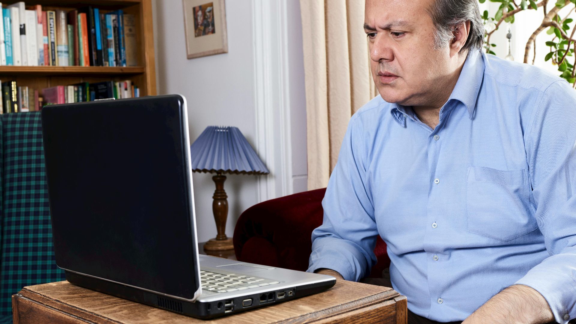 Middle-aged man focused on laptop in a cozy home library setting, surrounded by books.
