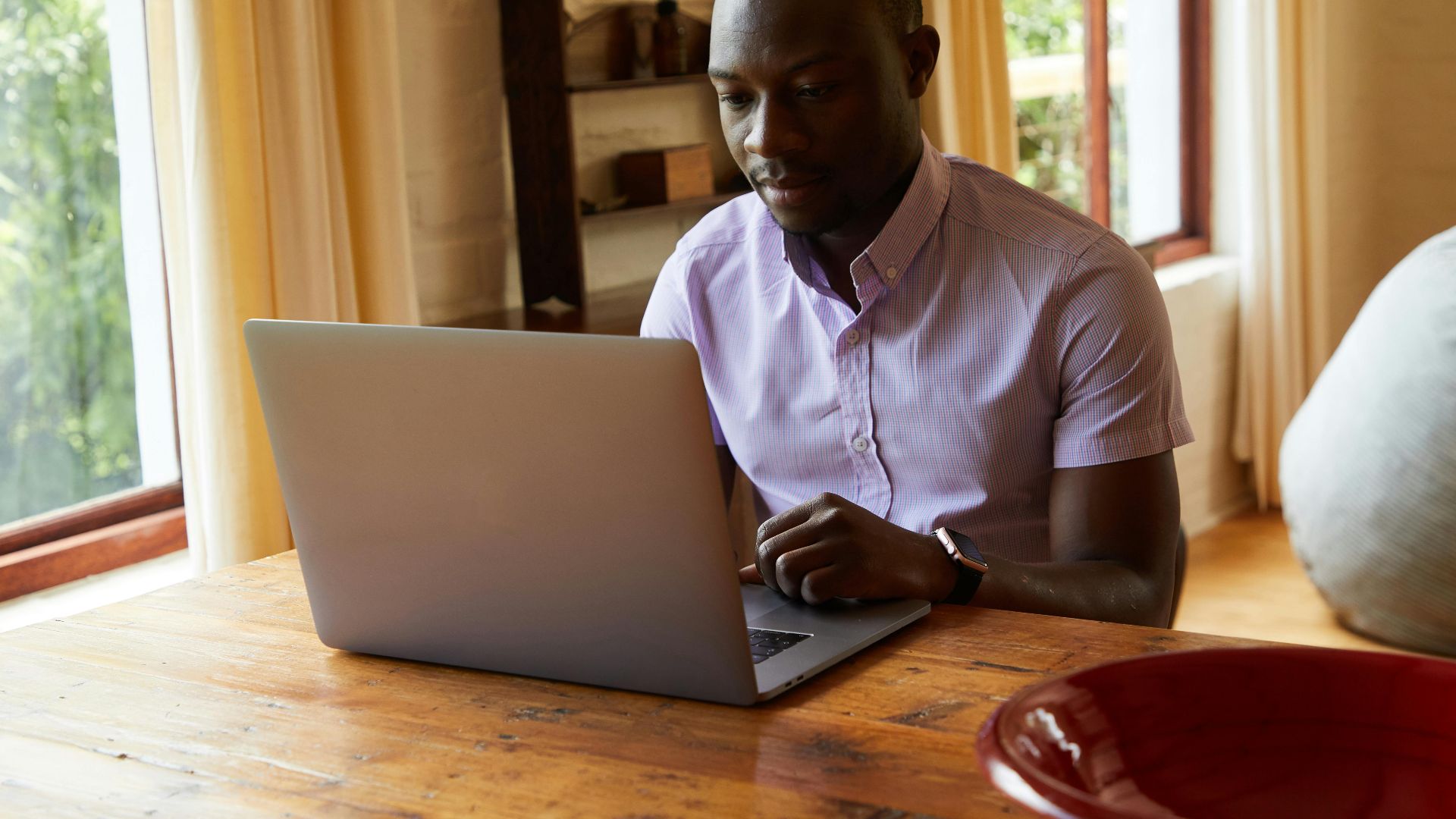 Serious African American male in shirt surfing internet on netbook placed on timber table in comfortable room