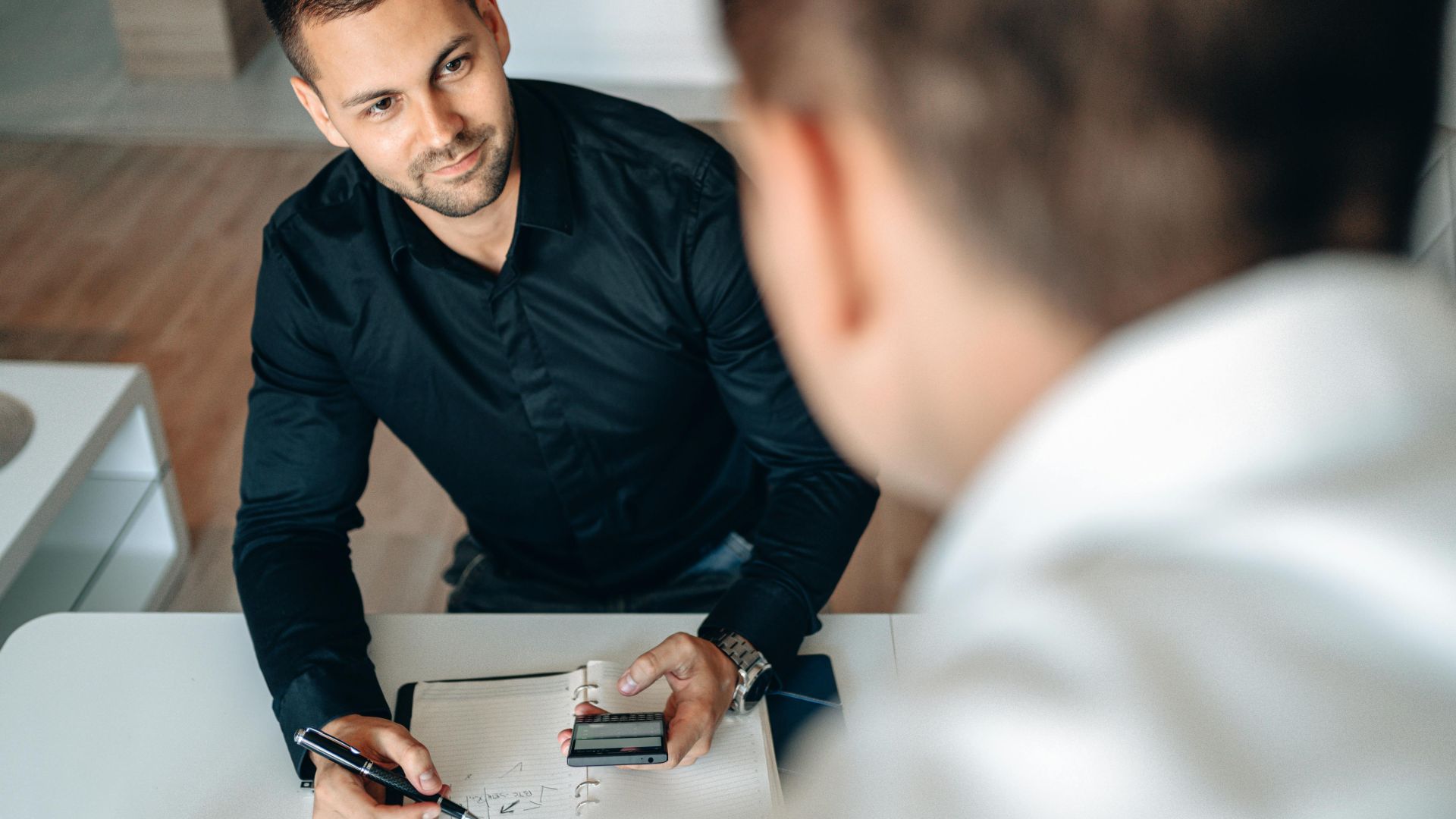 Two men in a business meeting indoors, discussing and taking notes.