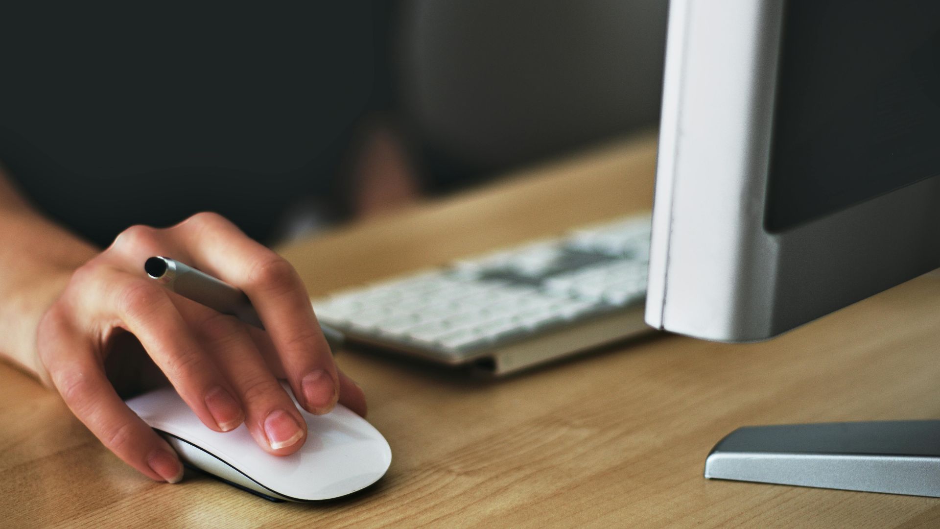 A hand using a wireless mouse at a modern desk setup with a computer and keyboard.