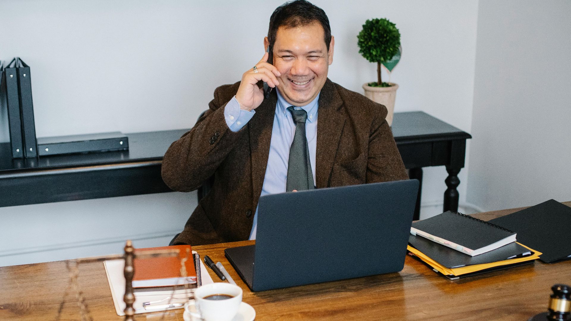 Joyful Asian male lawyer wearing formal suit having conversation on mobile phone and using laptop while working in modern law firm