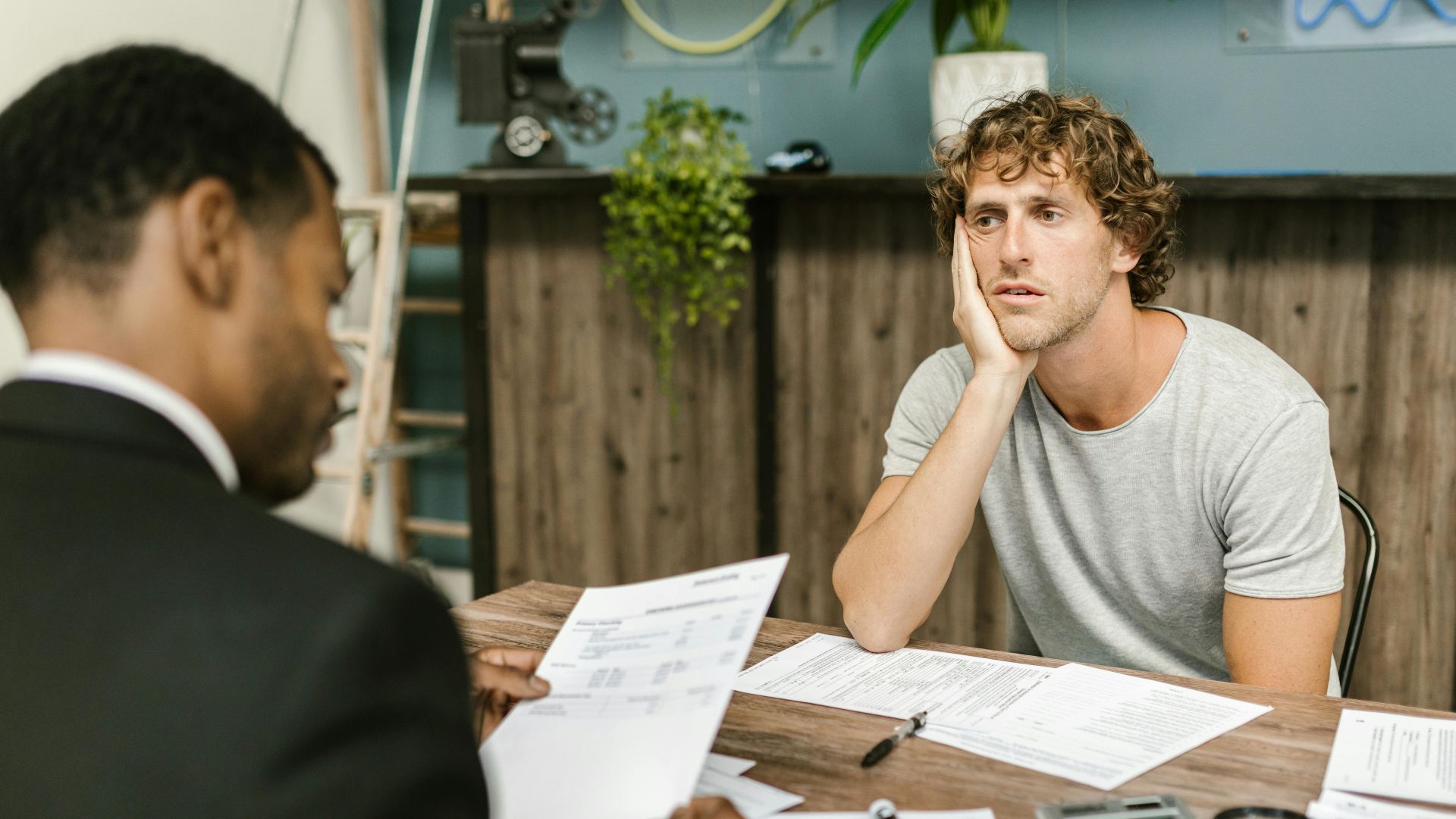 Man in office, appearing stressed during paperwork discussion, sitting opposite professional.