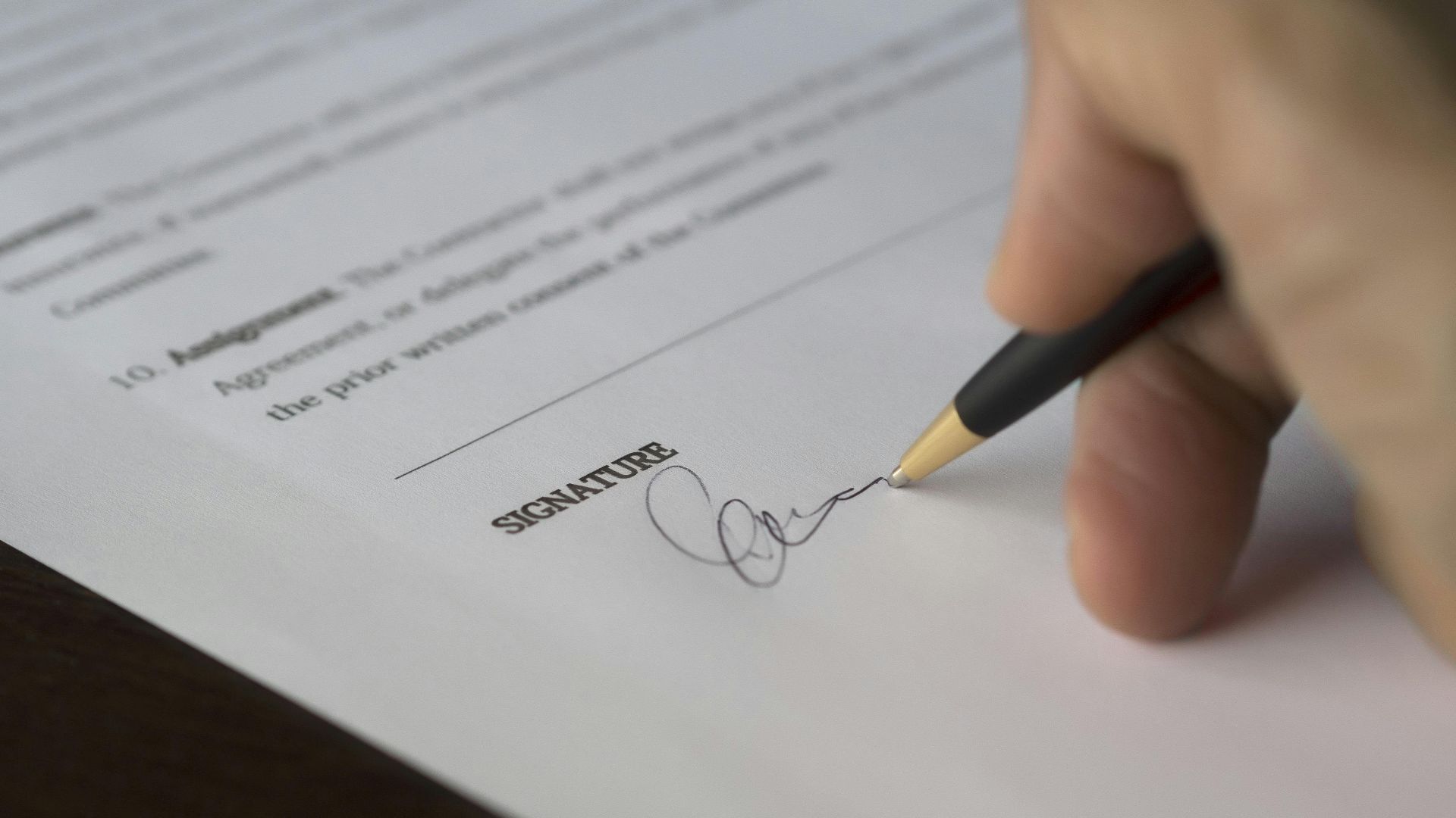 A hand signs a formal contract with a pen on a wooden desk.
