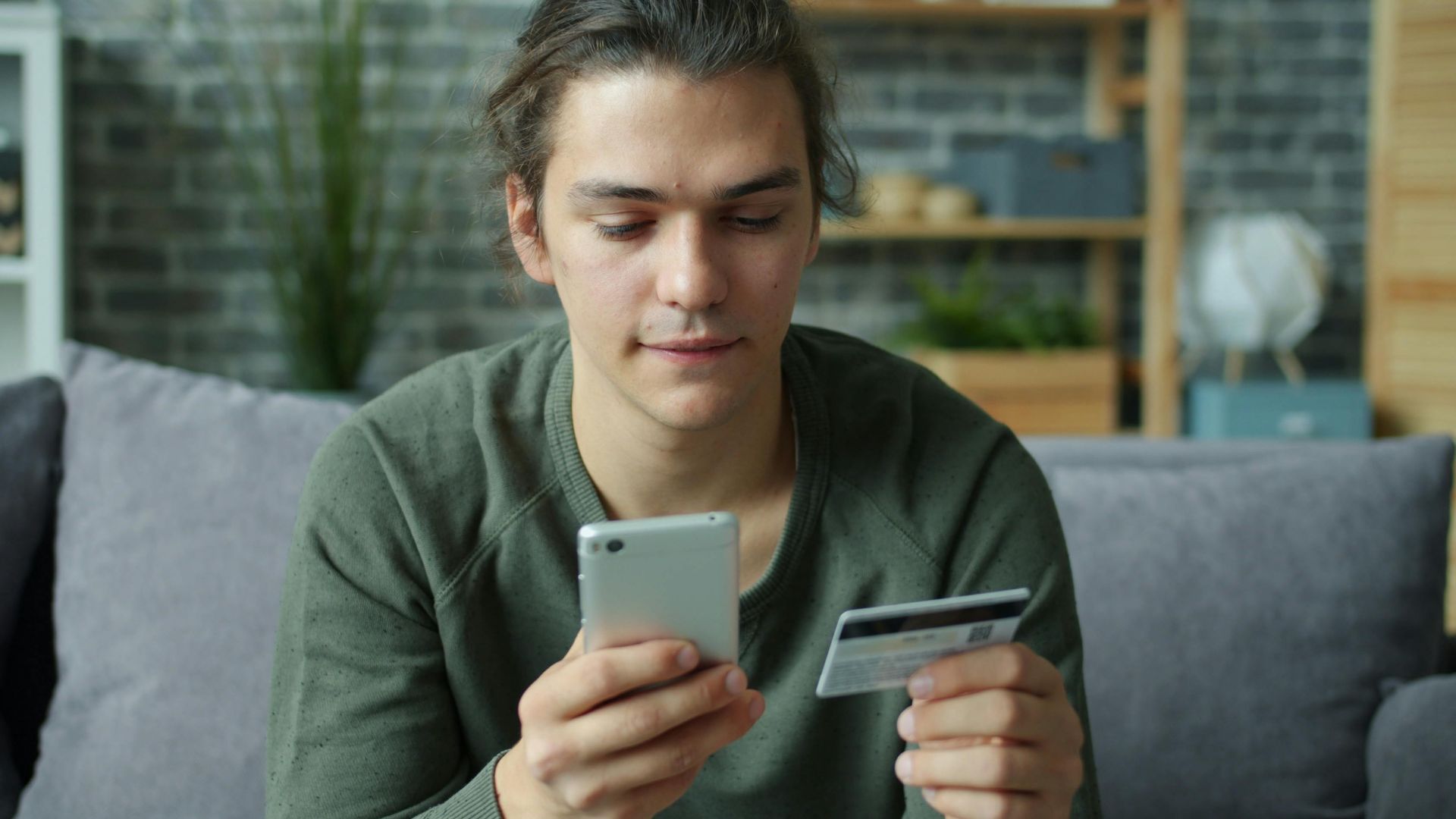 A young man using a smartphone and credit card for online shopping at home.