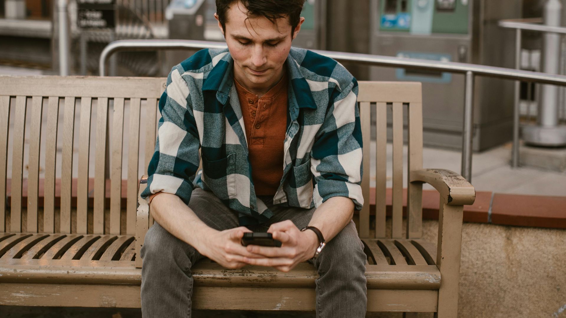 Man wearing plaid shirt using smartphone on a wooden bench at an urban train station.