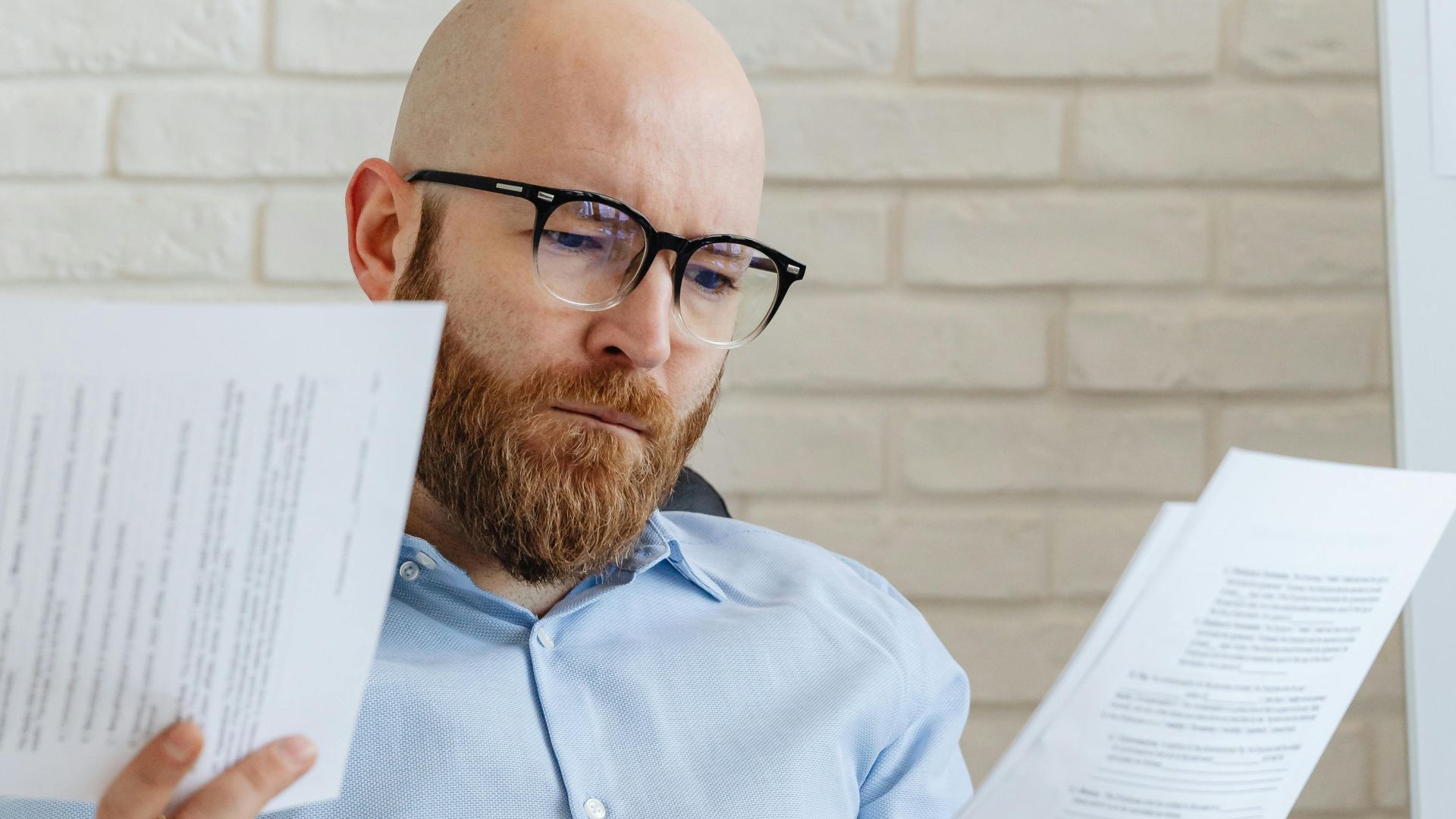 A serious-looking bald man with glasses reviews papers in an office against a brick wall.