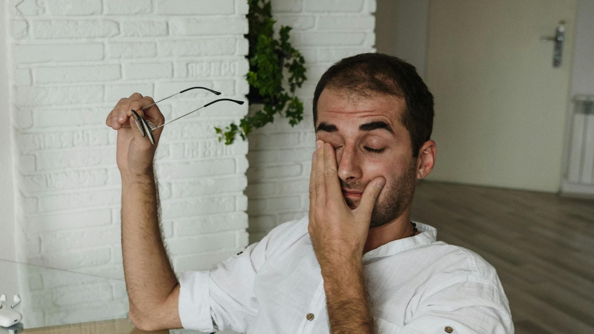 A tired man in a white shirt rests at a home office desk, holding eyeglasses and rubbing his eyes.
