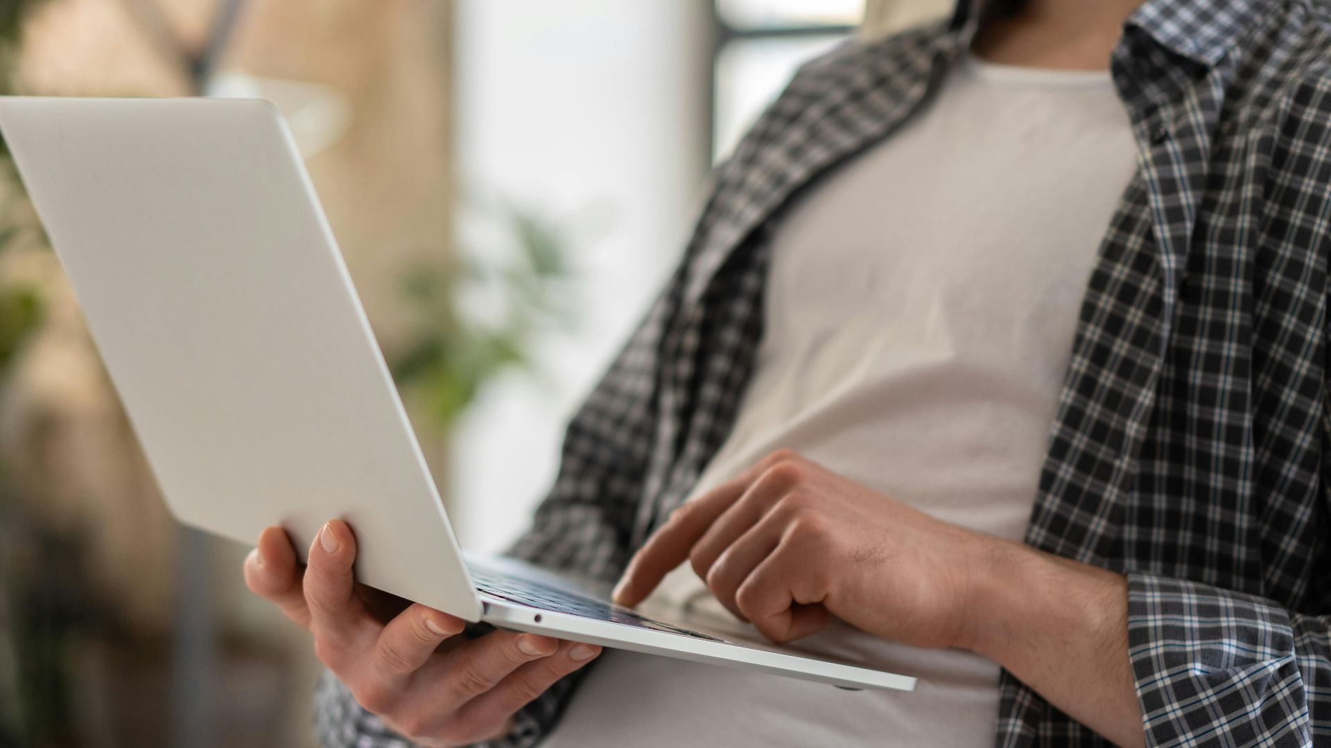 Young adult in casual shirt using a laptop indoors with blurred background and shallow focus.