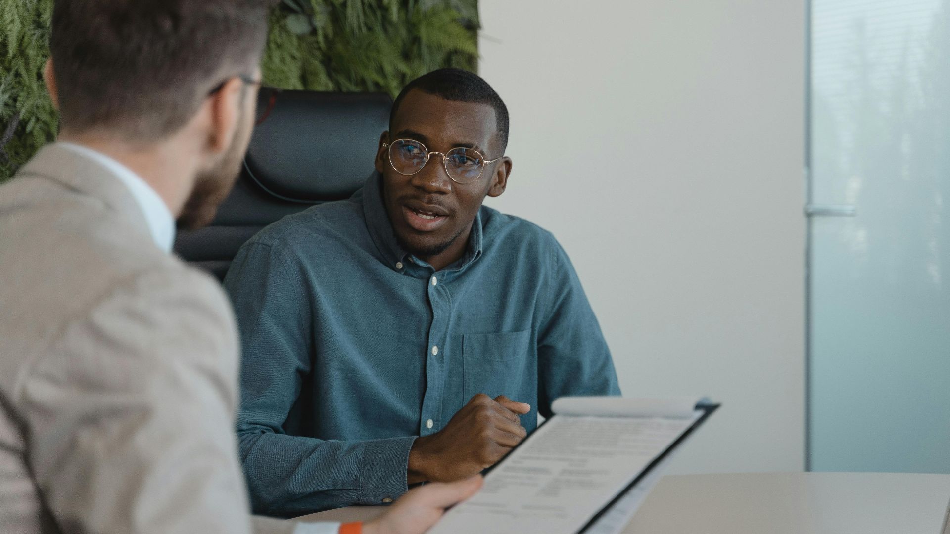 Two men engaged in a professional meeting at an office table with documents and a plant wall background.