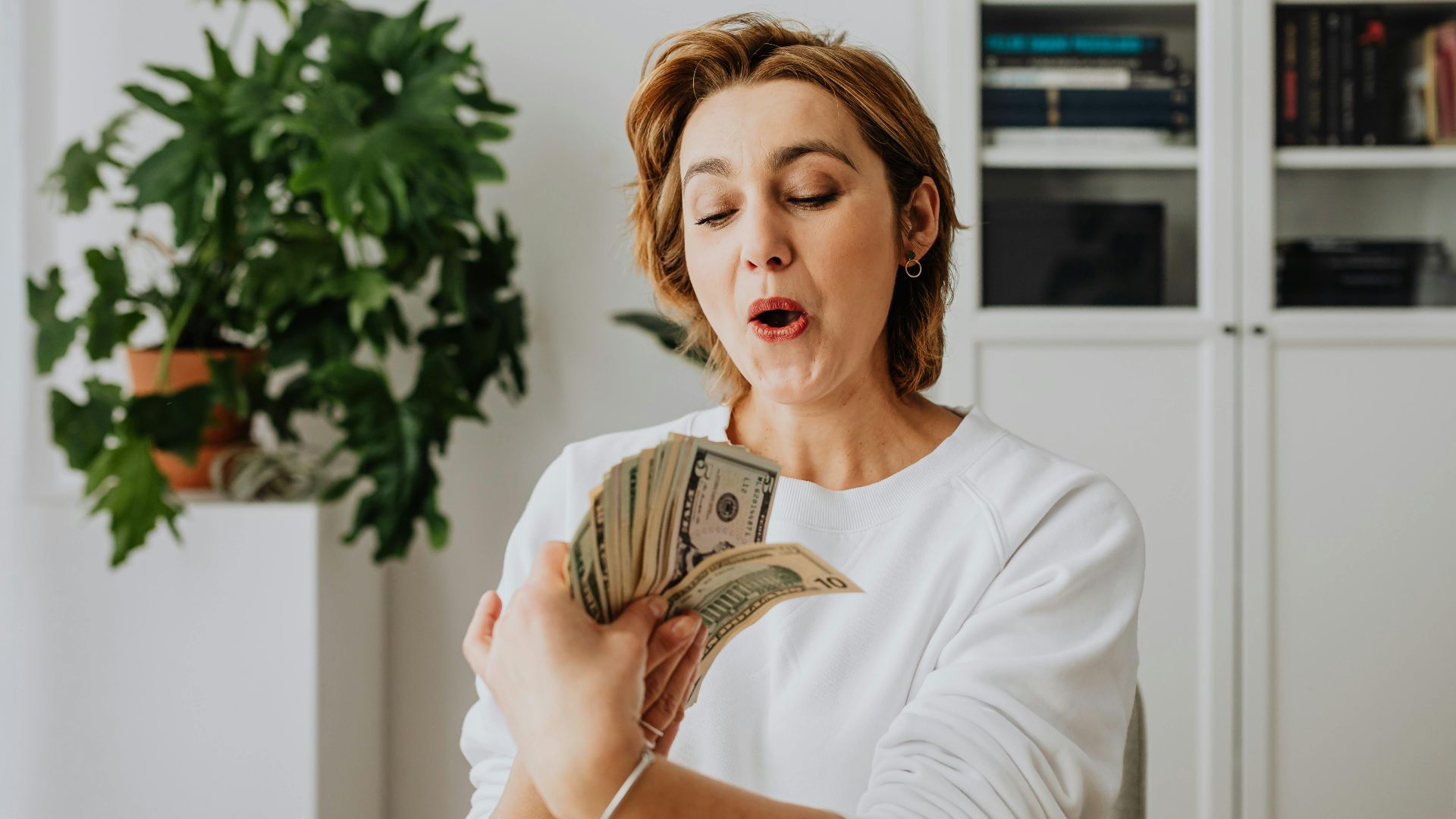A woman in a white sweater excitedly counts American dollar bills indoors, reflecting joy and prosperity.