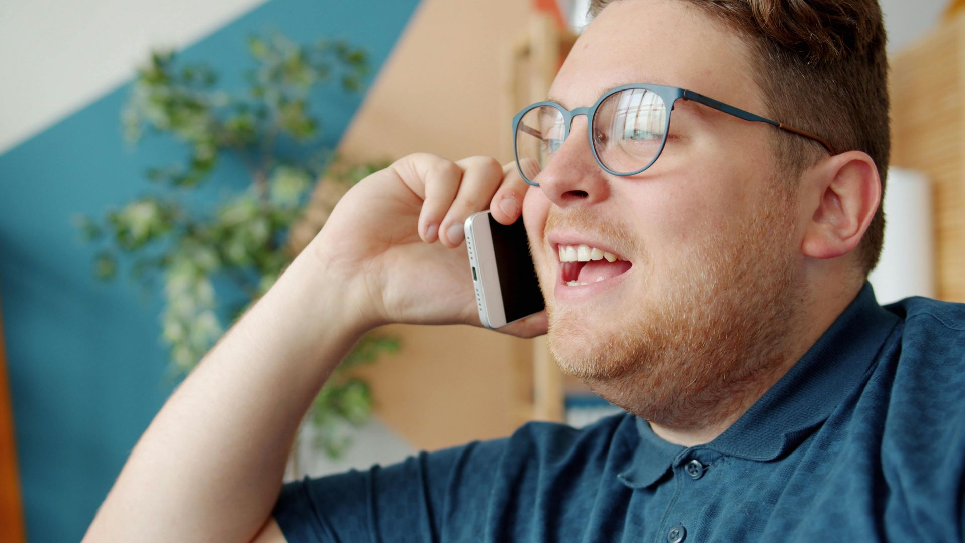 Happy adult man with glasses having a phone conversation inside a bright room.