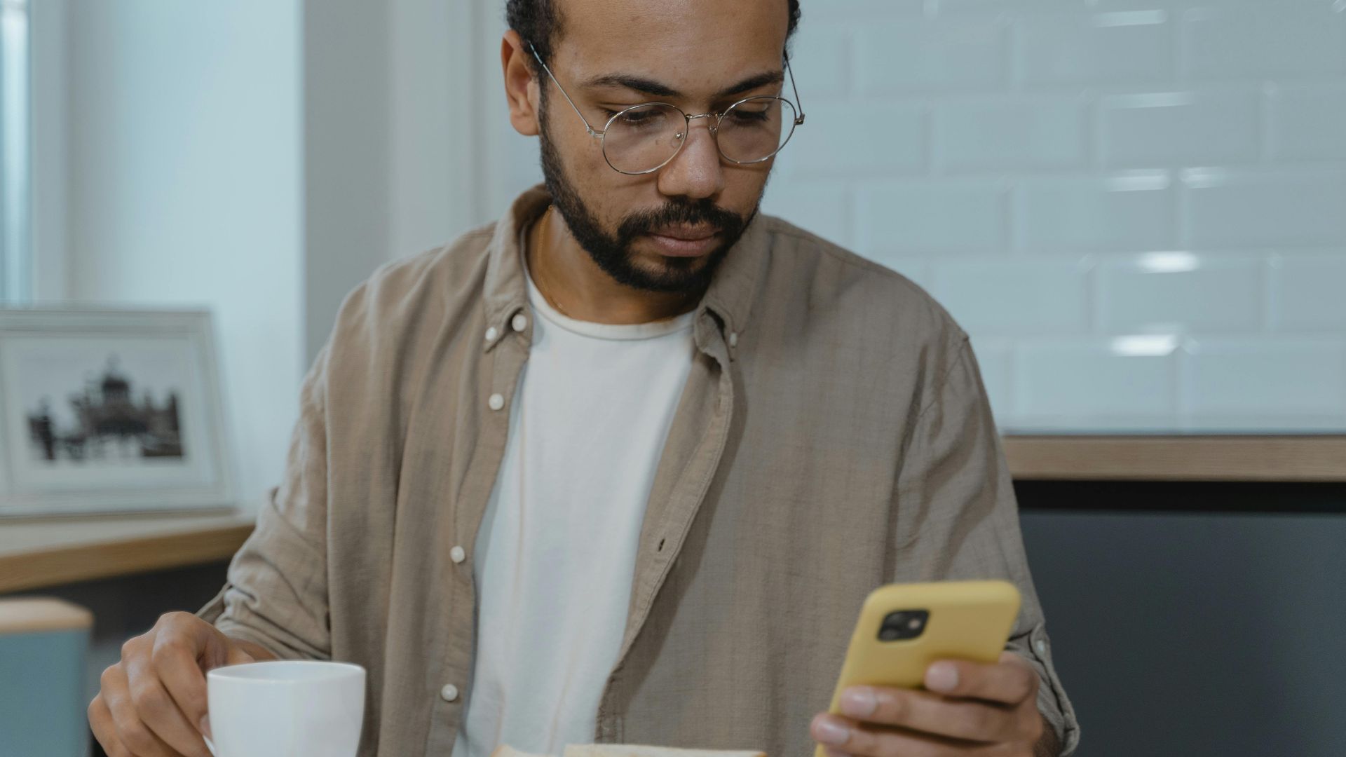 A man with eyeglasses using a smartphone while enjoying breakfast indoors. Perfect for lifestyle and technology themes.