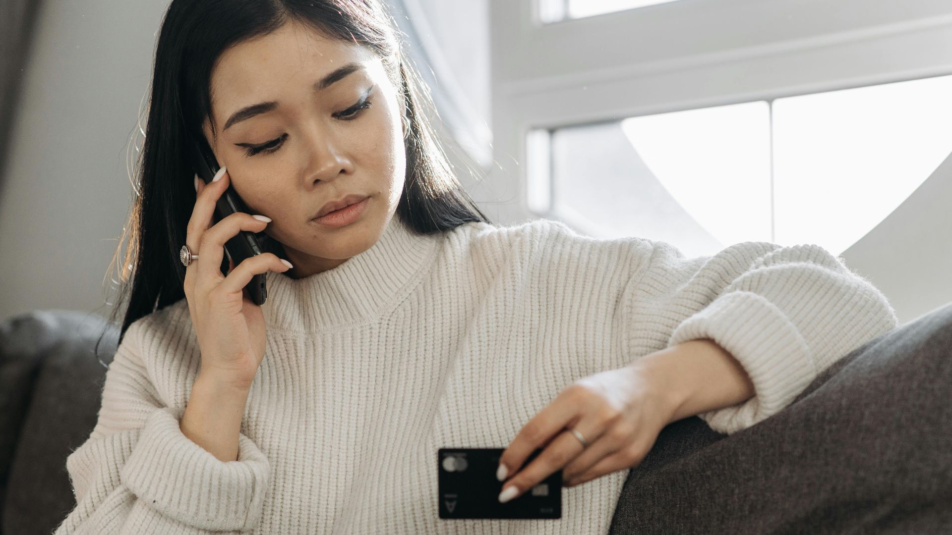 A woman sits on a sofa, multitasking with a phone and credit card, focusing on online shopping.