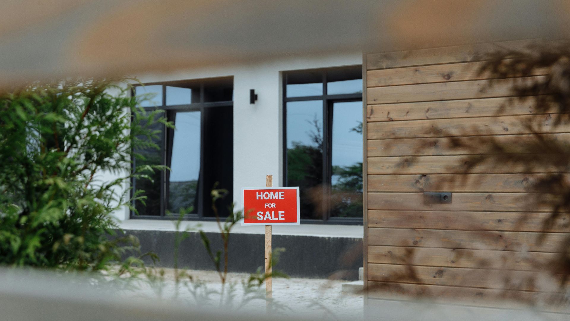 A contemporary home with a 'For Sale' sign in the front yard, framed by greenery.
