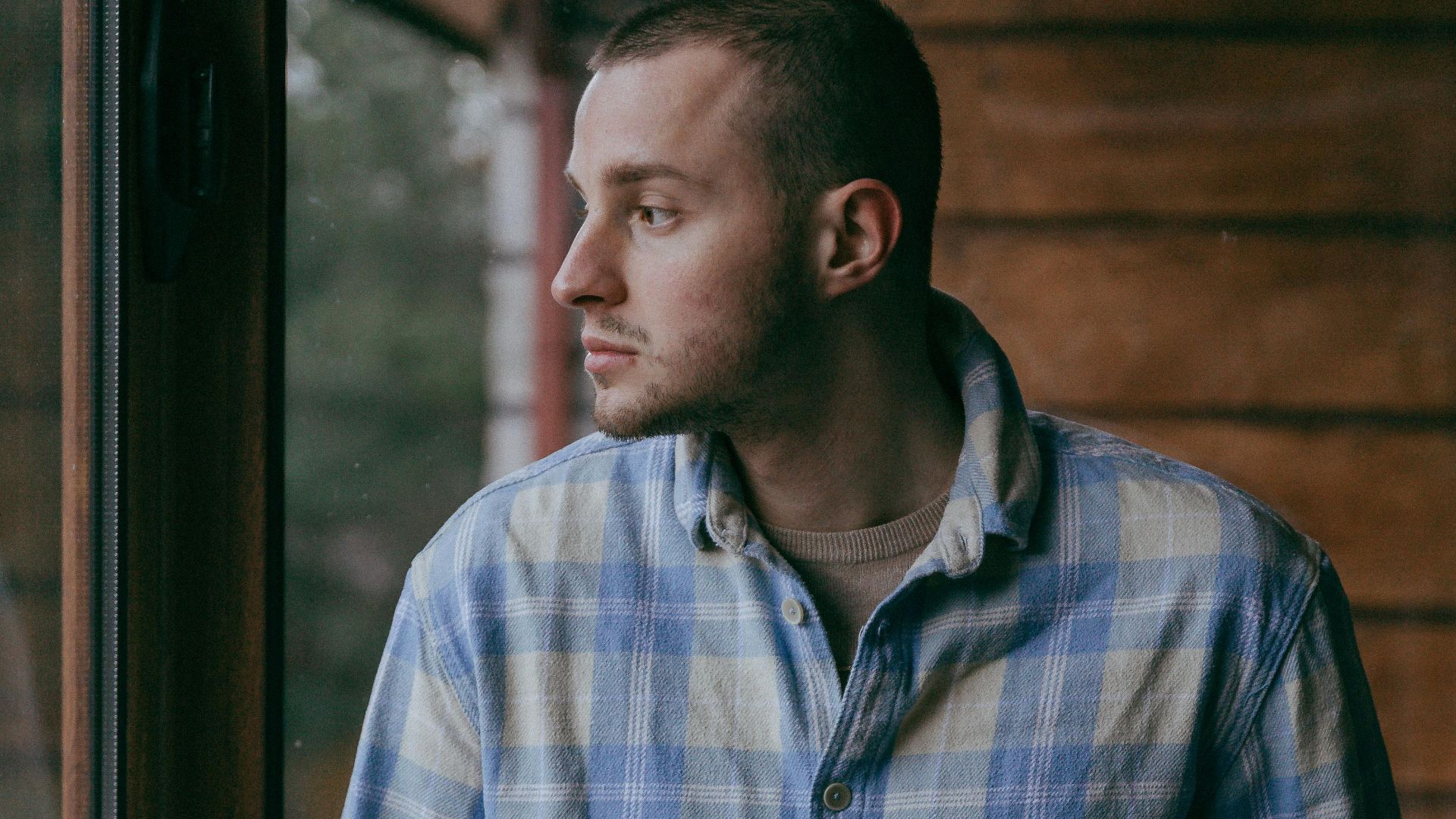 Young man in casual checkered shirt gazing thoughtfully out a window.
