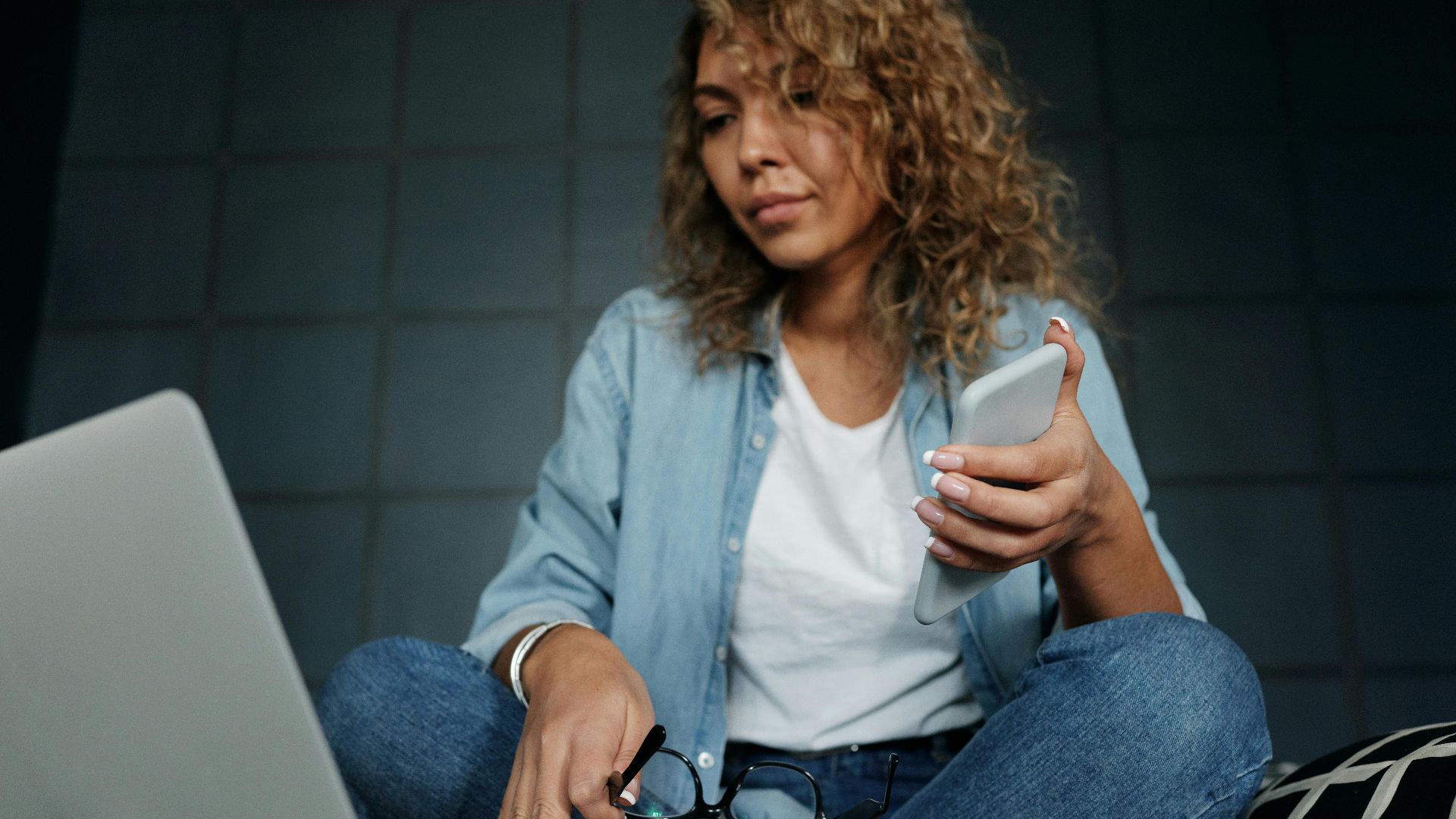 A woman comfortably working from home on a laptop and smartphone, sitting on bed.