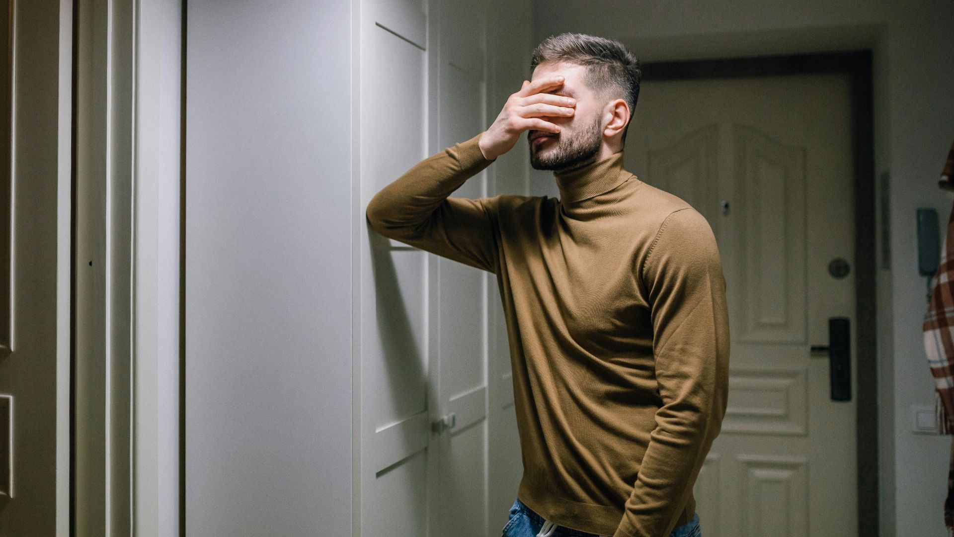 A man wearing a brown sweater stands indoors, appearing distressed with a hand on his face.