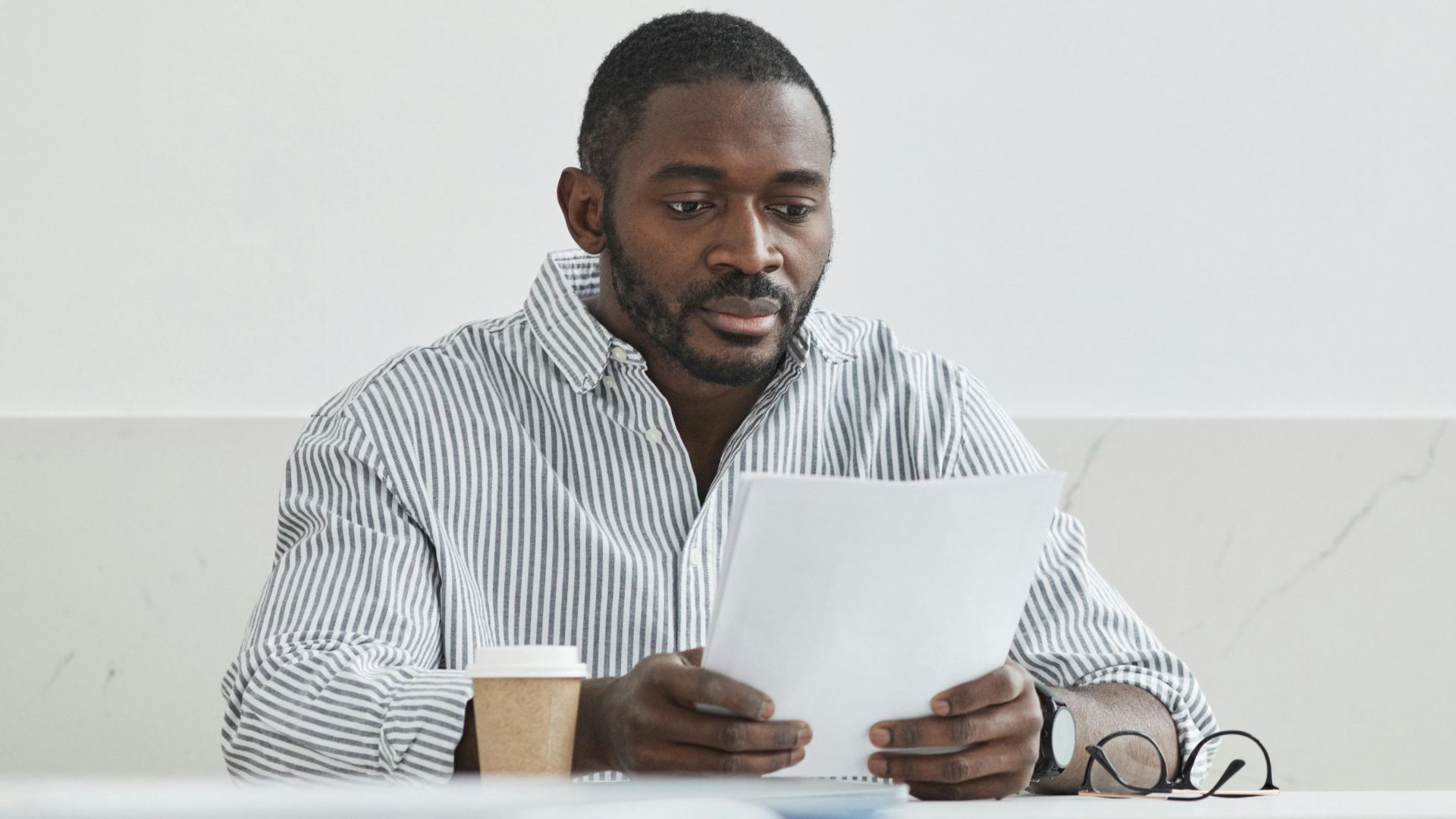 African American man sitting indoors, reading papers with a coffee cup nearby.