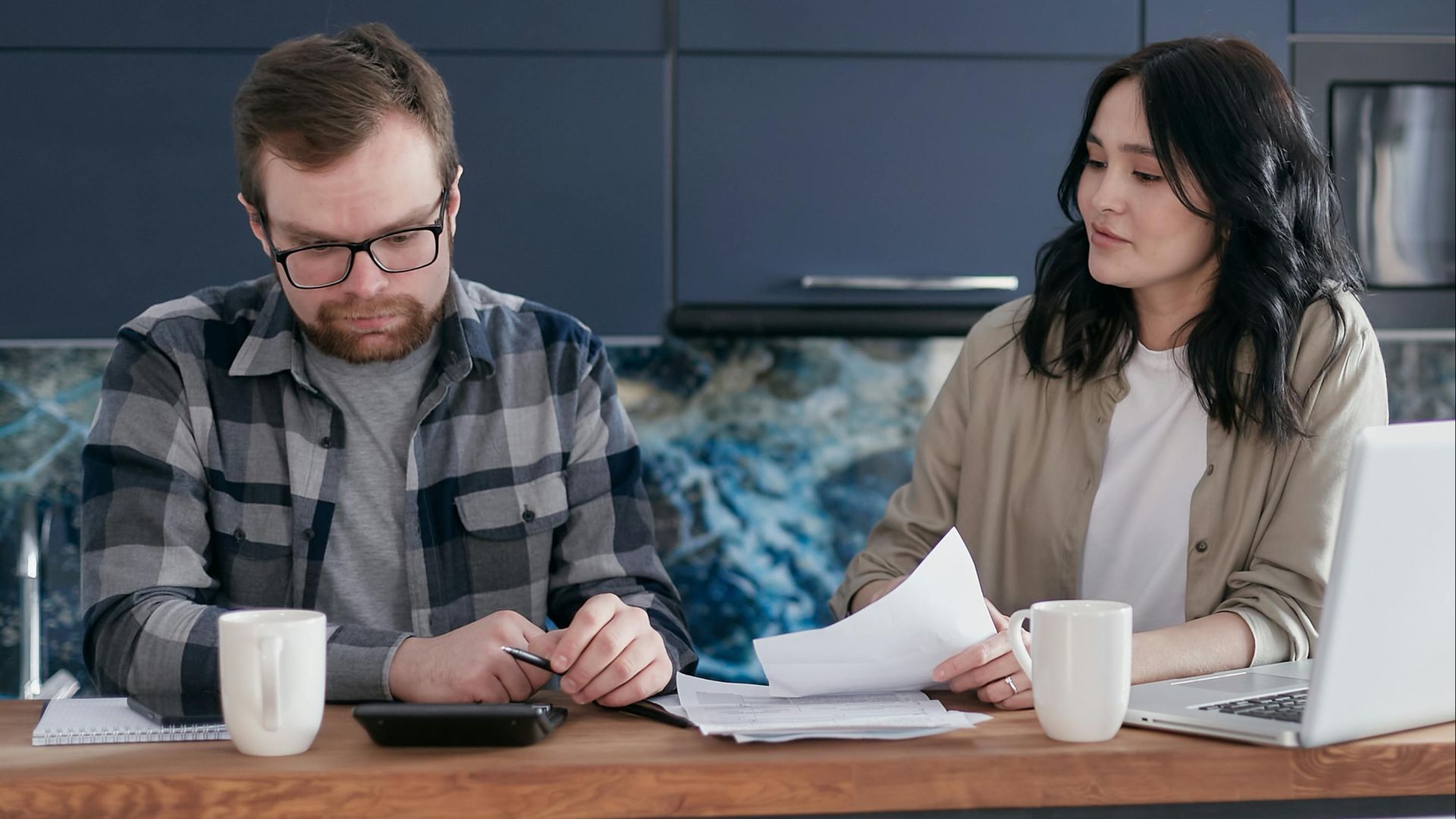 A couple discusses bills and finances using a laptop and calculator in their modern kitchen.