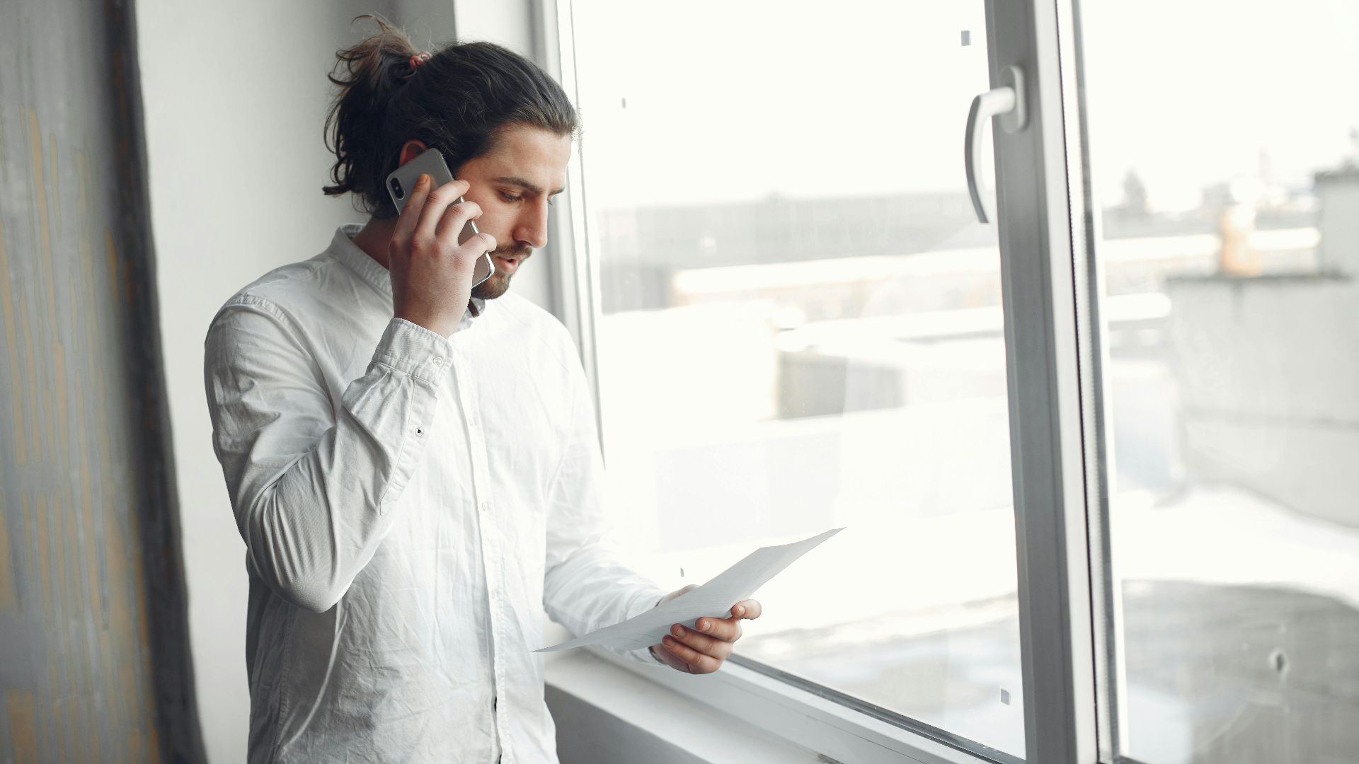 Young man in white shirt, on phone call holding a document, standing by a large window.