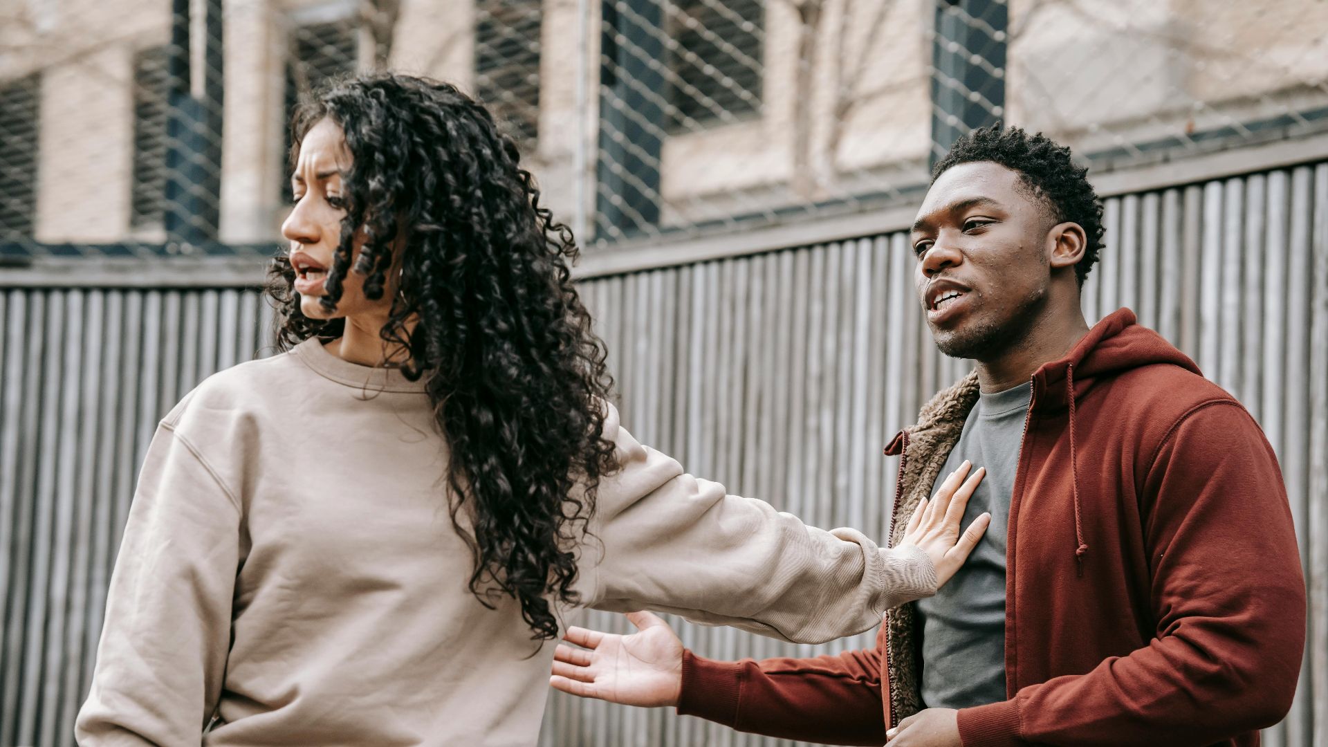 A couple in casual clothing appears to be in a disagreement outdoors with grey fencing background.