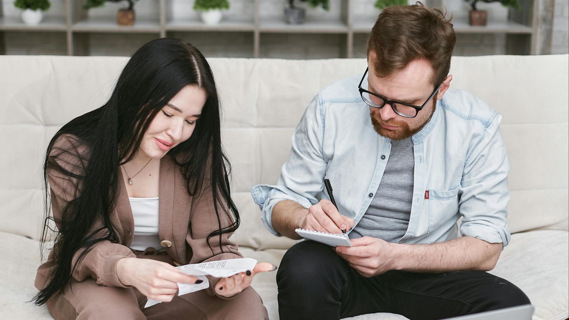A man and woman sitting indoors, reviewing expenses with notebooks and money on the table.