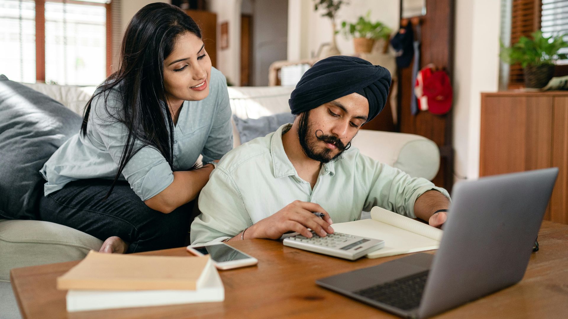 Smiling focused ethnic woman sitting on sofa at home watching working ethnic husband calculating sitting at coffee table among calculator and laptop and smartphone and books and notebook in living room