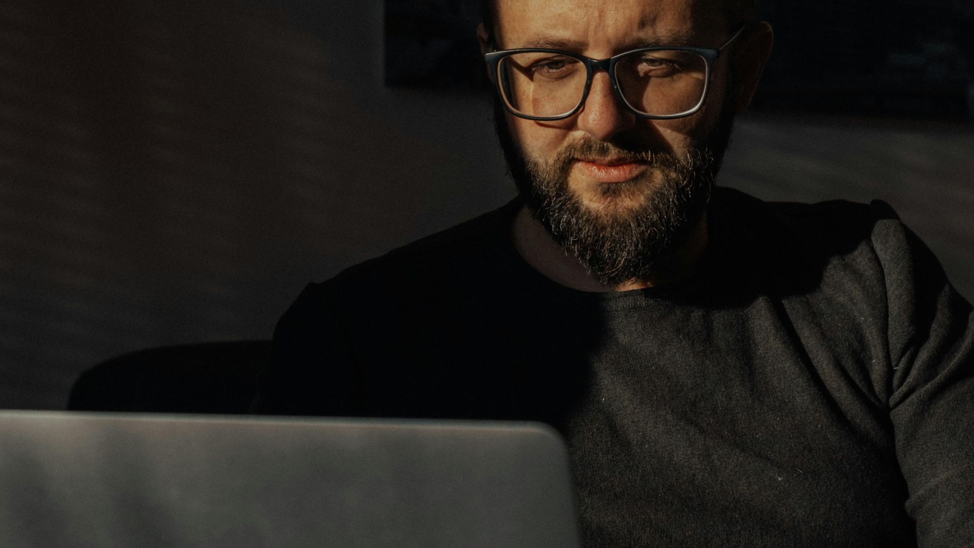 Bearded man with eyeglasses working on a laptop in a dimly lit room, focusing intently.