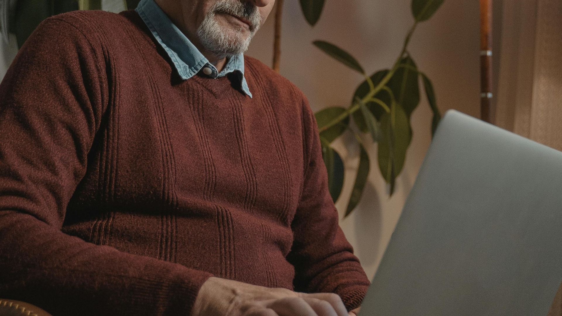 Senior adult working remotely on a laptop, seated in a cozy indoor setting with warm lighting.
