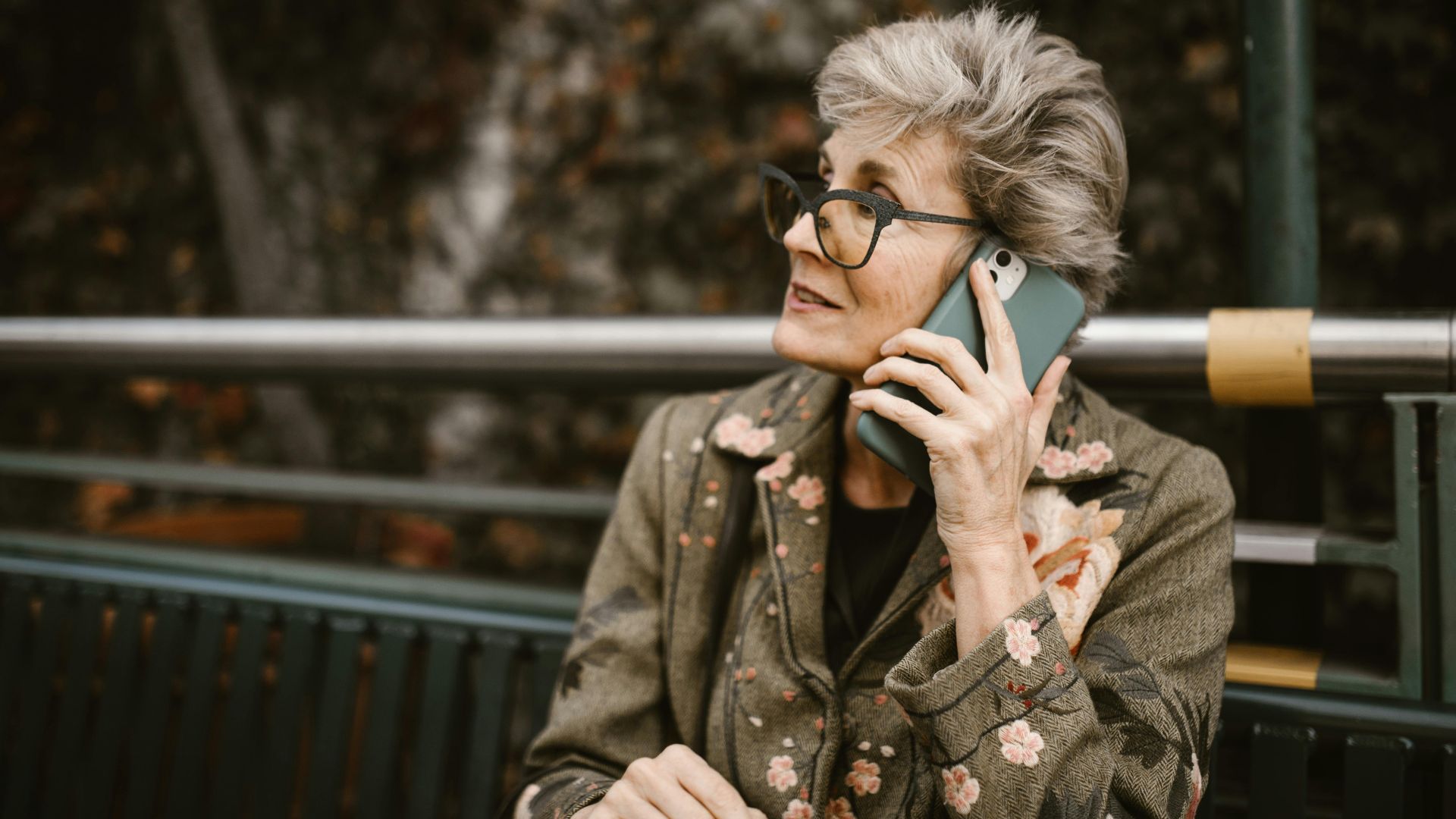 Elderly woman with glasses talking on a smartphone while seated outdoors.