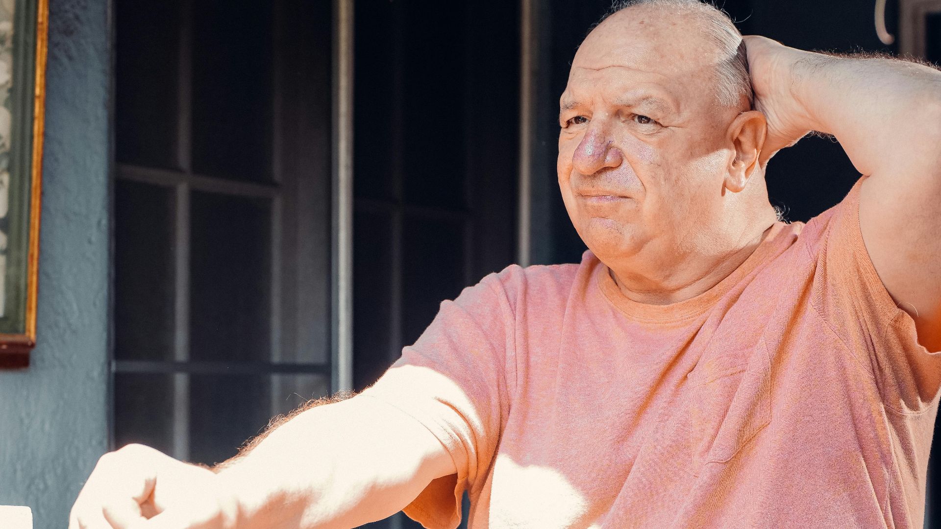 Portrait of a senior man in an orange shirt, reflecting outdoors, hand on head in thought.