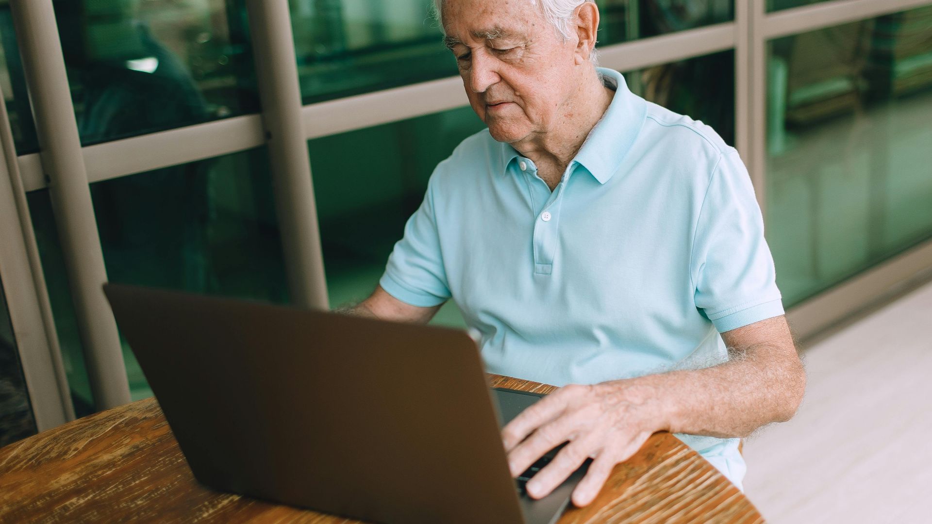 Elderly man sitting at a wooden table, working on a laptop indoors.