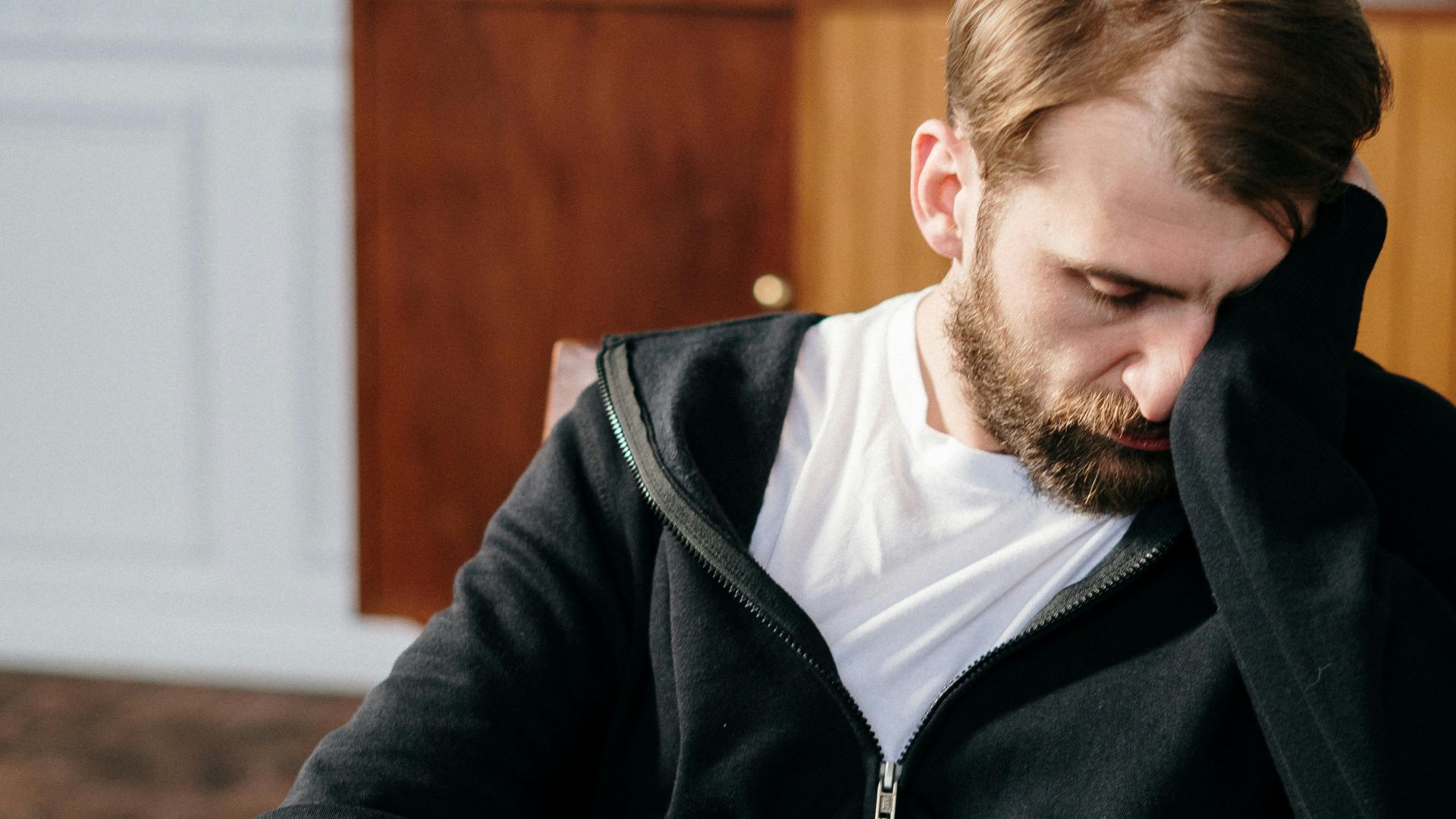 Bearded man in a black hoodie sitting in a chair indoors, appearing deep in thought.