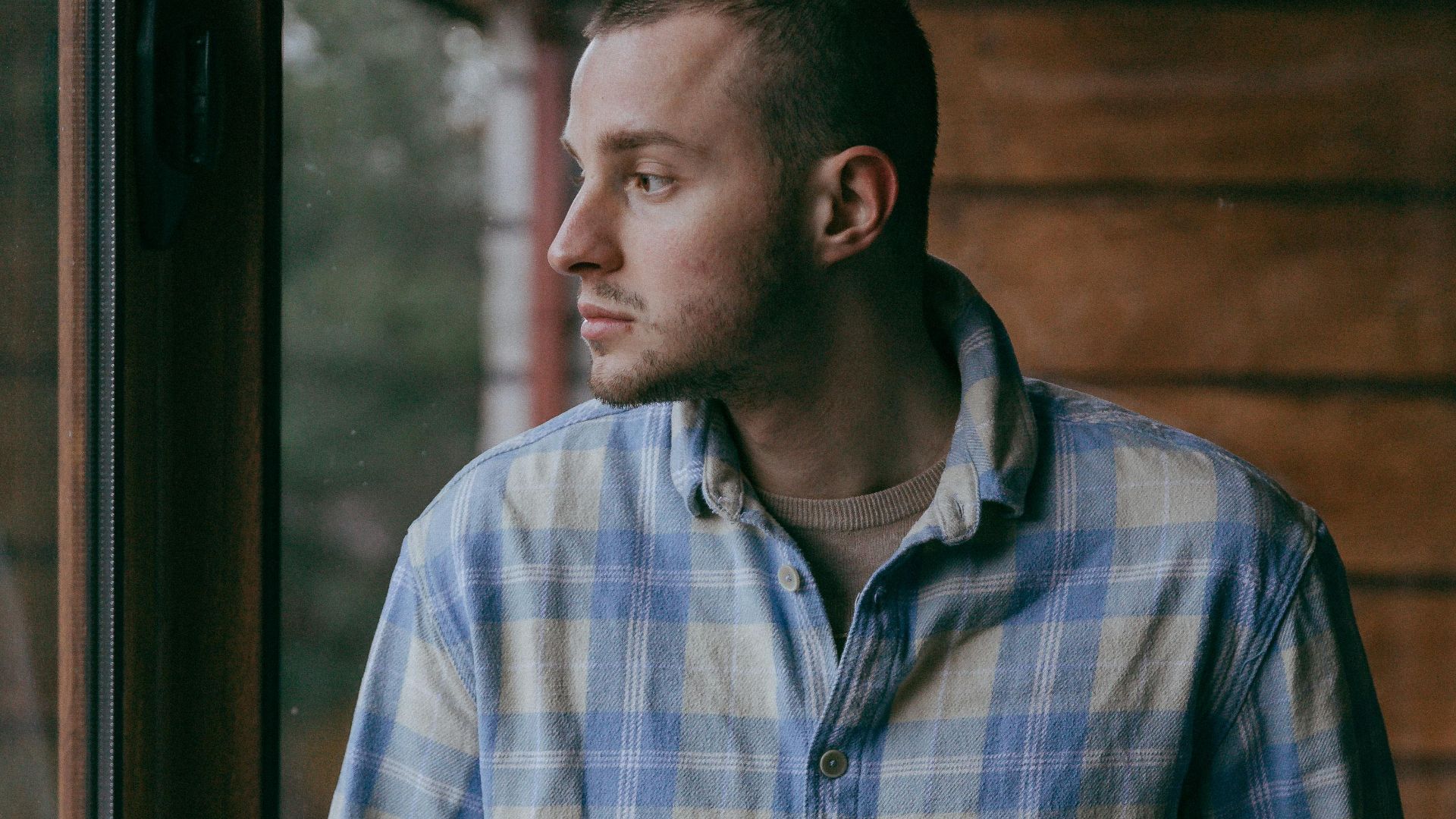 Young man in casual checkered shirt gazing thoughtfully out a window.