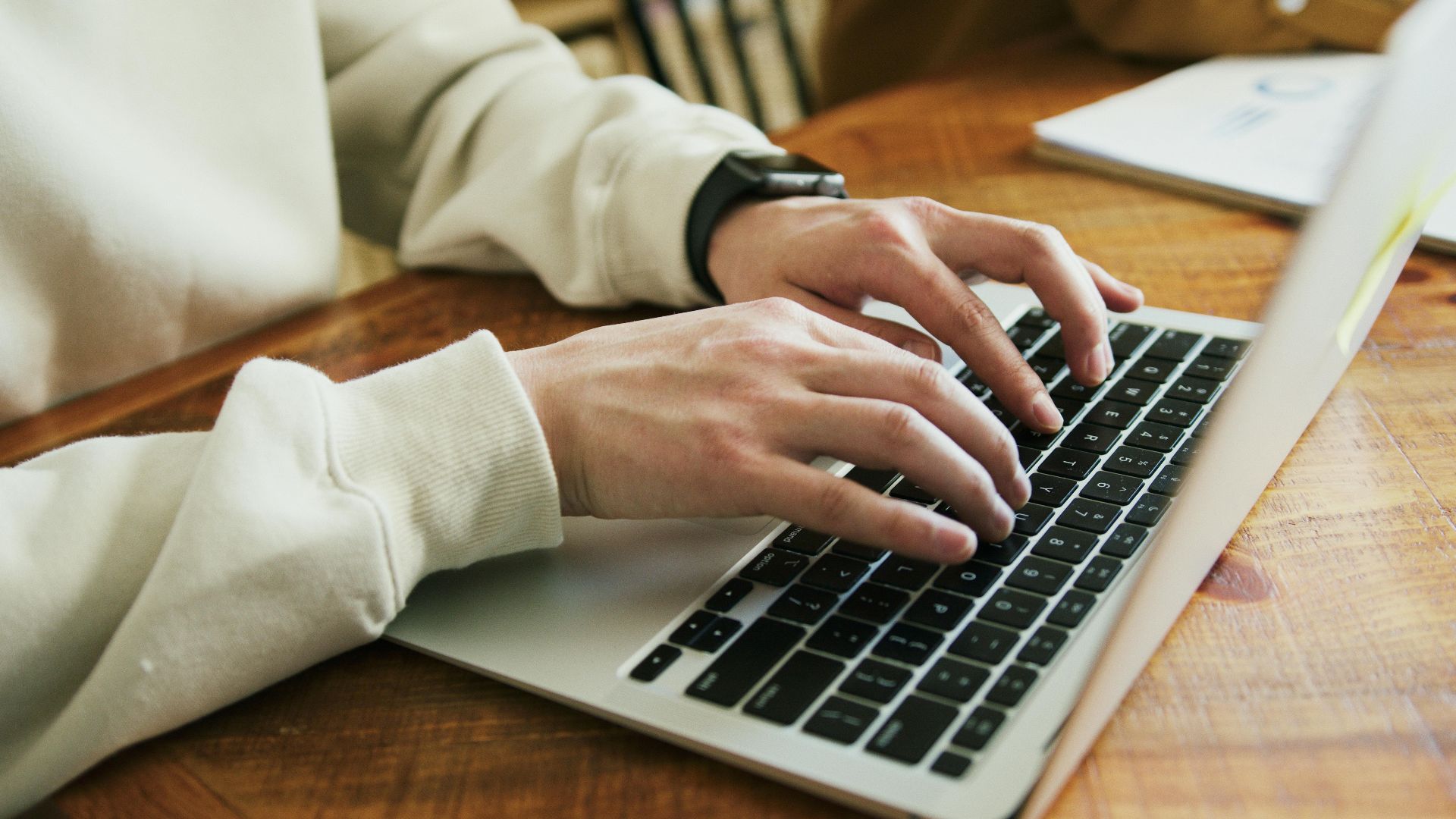 Hands typing on a laptop keyboard on a wooden desk, close-up view.