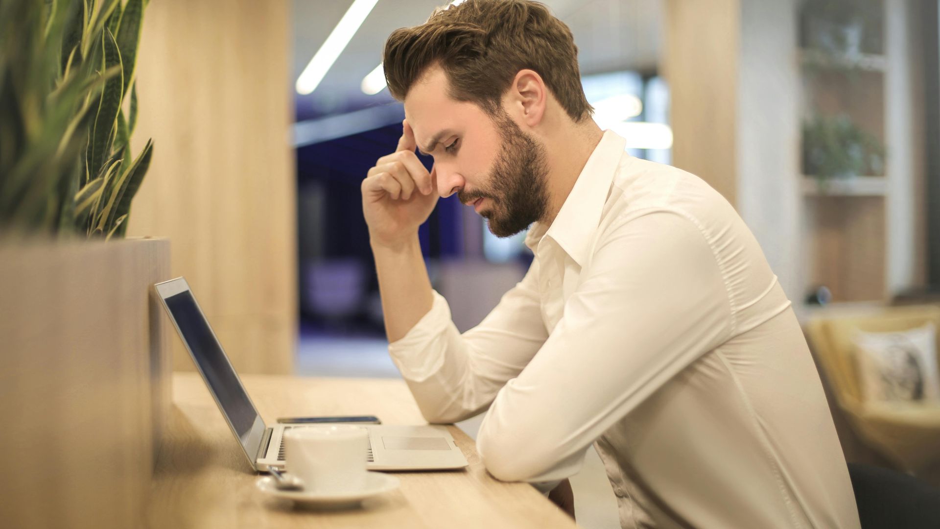 A thoughtful man in a shirt works on his laptop at a modern indoor office space.