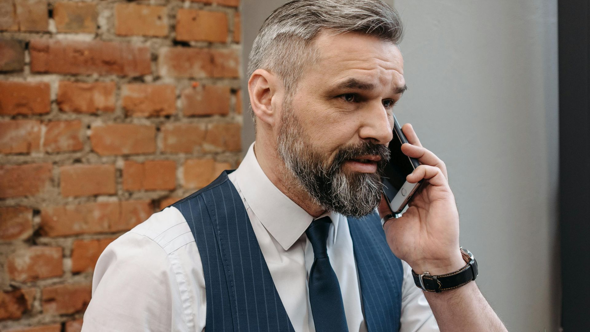 A stylish bearded man in formal attire talking on a smartphone in an office setting with exposed brick.