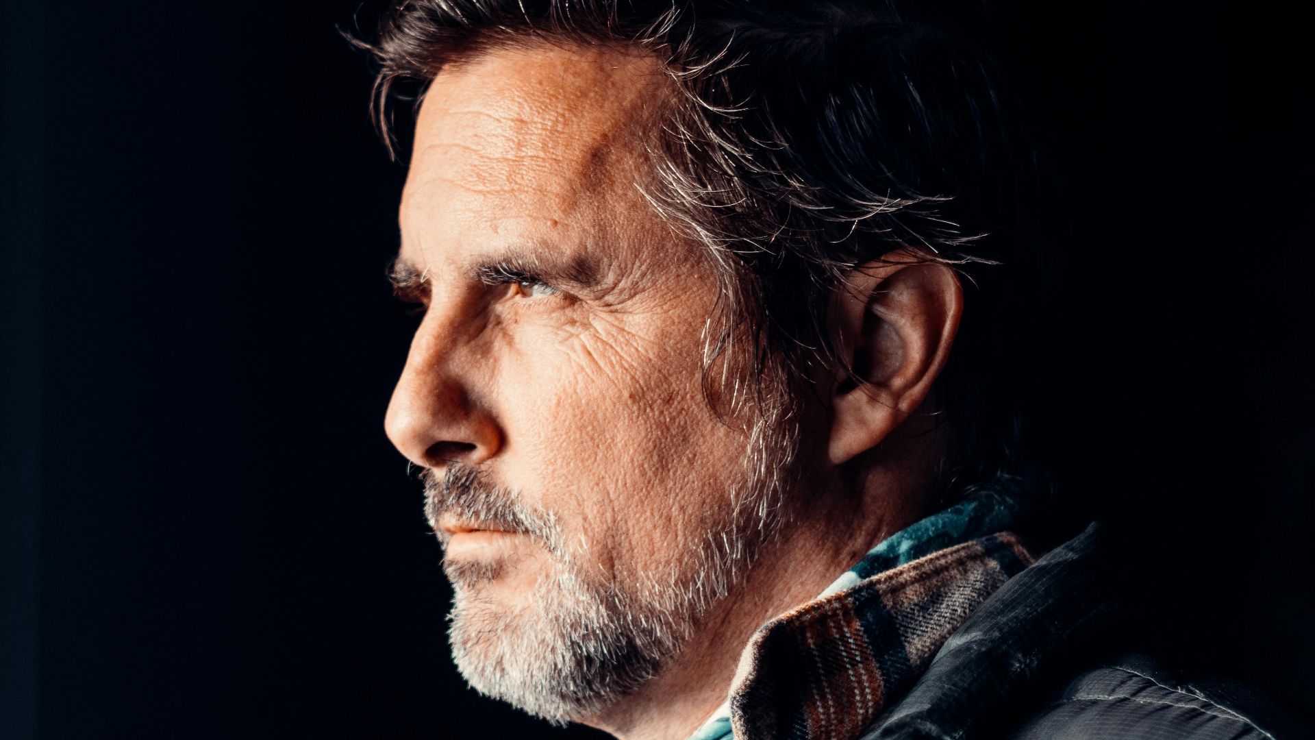 An elderly man with facial hair gazes pensively out a window in a dimly lit room.