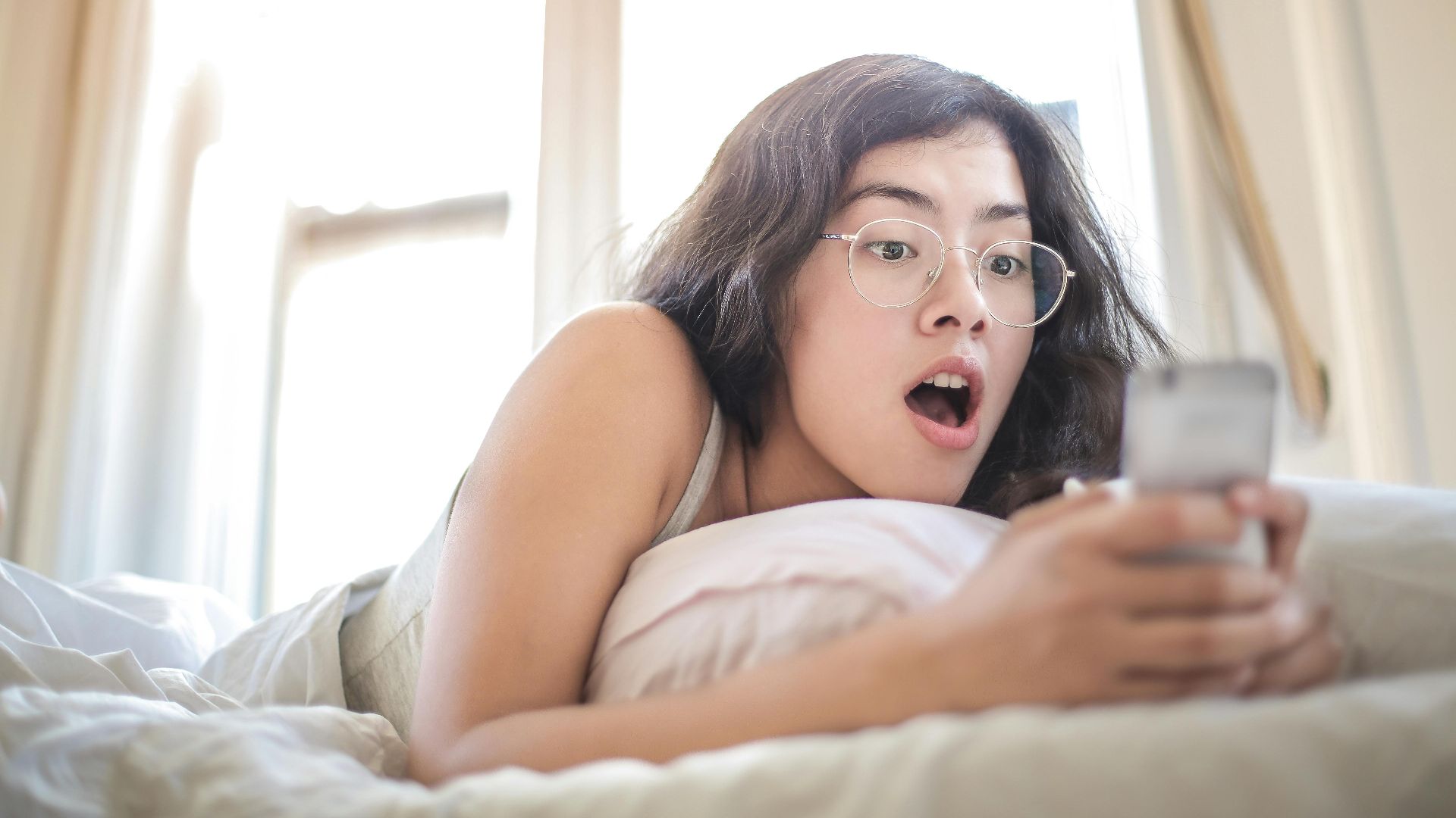 Young woman lying on bed, surprised by something on her smartphone, in a bright bedroom setting.