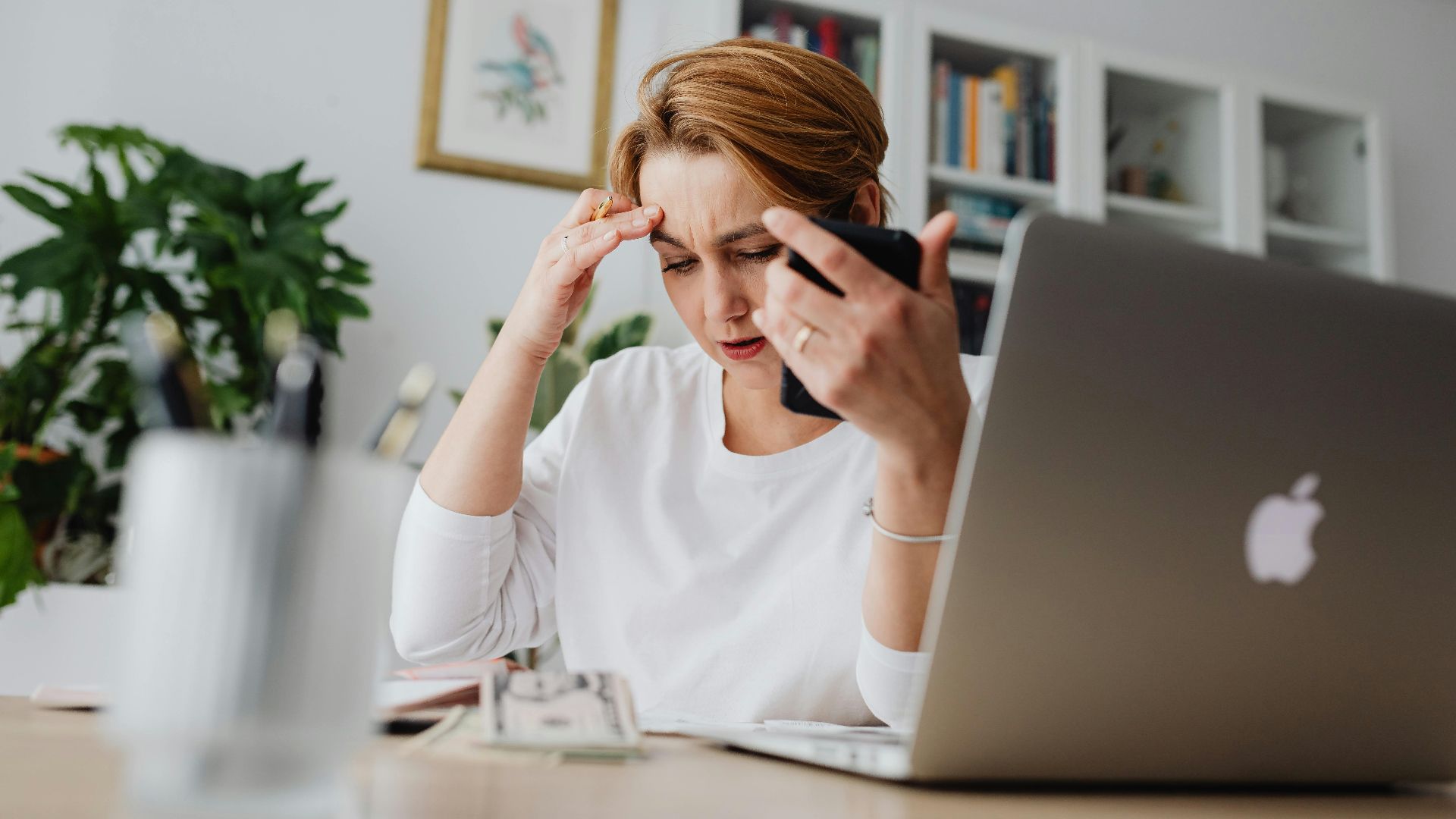 A stressed woman holds her phone, working at a desk with laptop and papers.