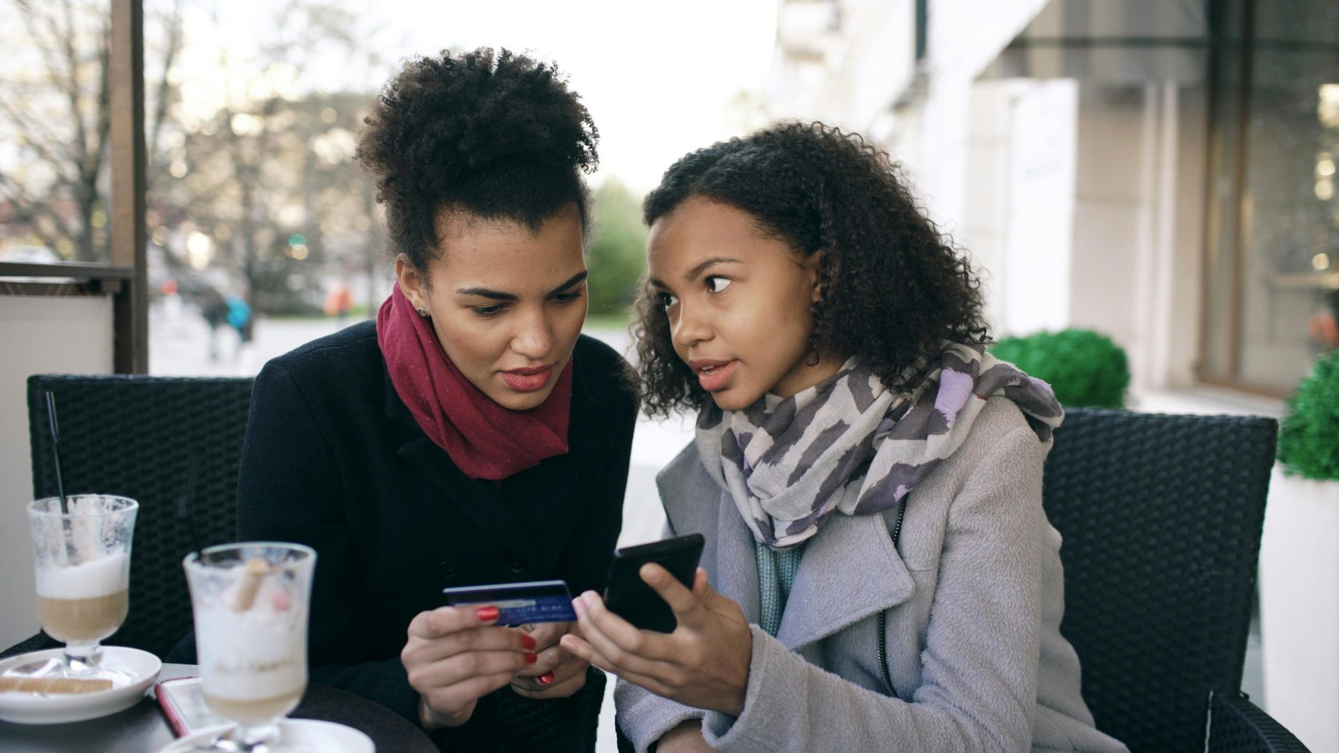 Two women shopping online with a smartphone and credit card at an outdoor cafe.